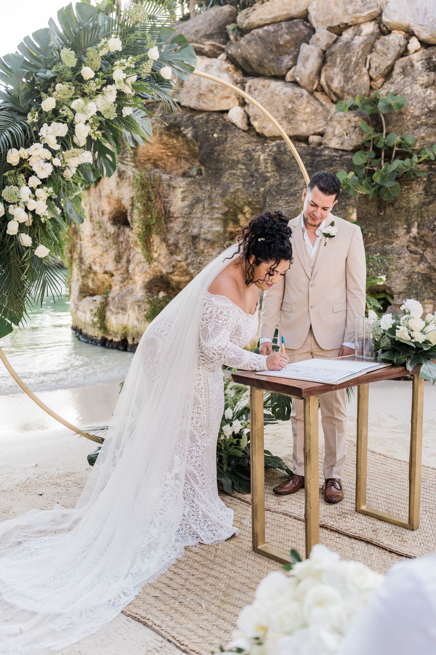 Xcaret Arte Wedding - Bride signing wedding certificate at beach ceremony with floral arch, groom watching, tropical setting.