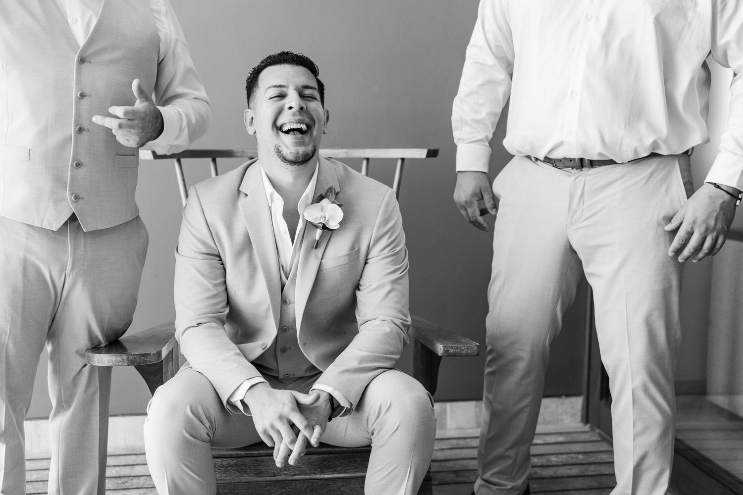 Xcaret Arte Wedding - Groom laughing joyfully while seated, surrounded by groomsmen in light suits, black and white wedding photo.