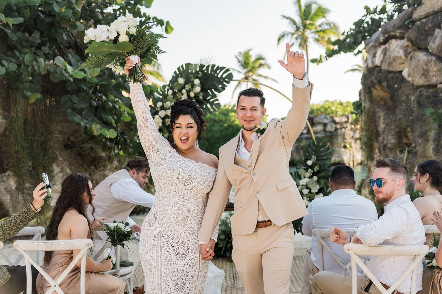 Xcaret Arte Wedding - Joyful newlyweds celebrate after outdoor wedding ceremony, bride in lace gown and groom in beige suit, tropical venue.