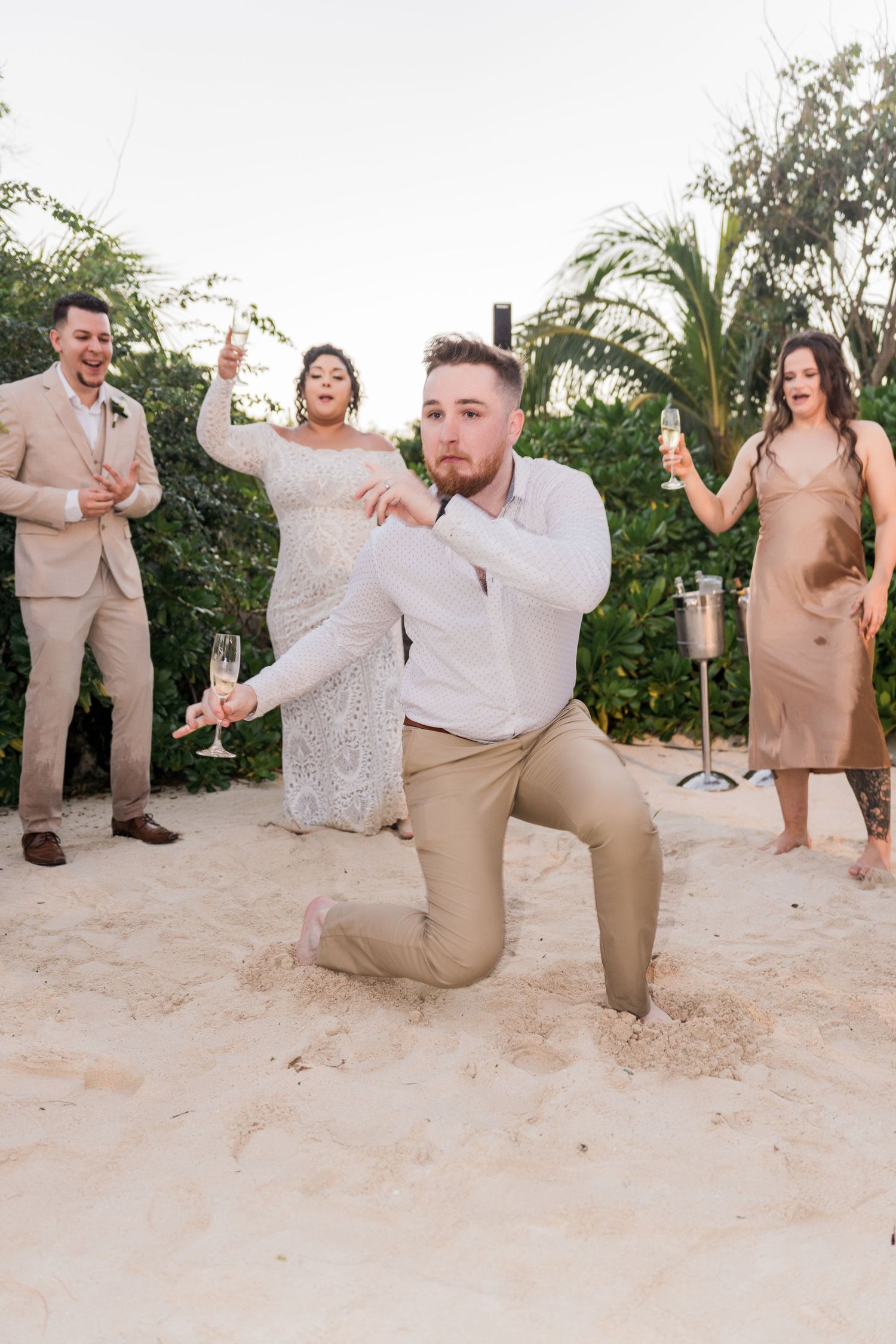 Xcaret Arte Wedding - Groom kneels on sandy beach while bride and wedding party celebrate with champagne glasses outdoors.