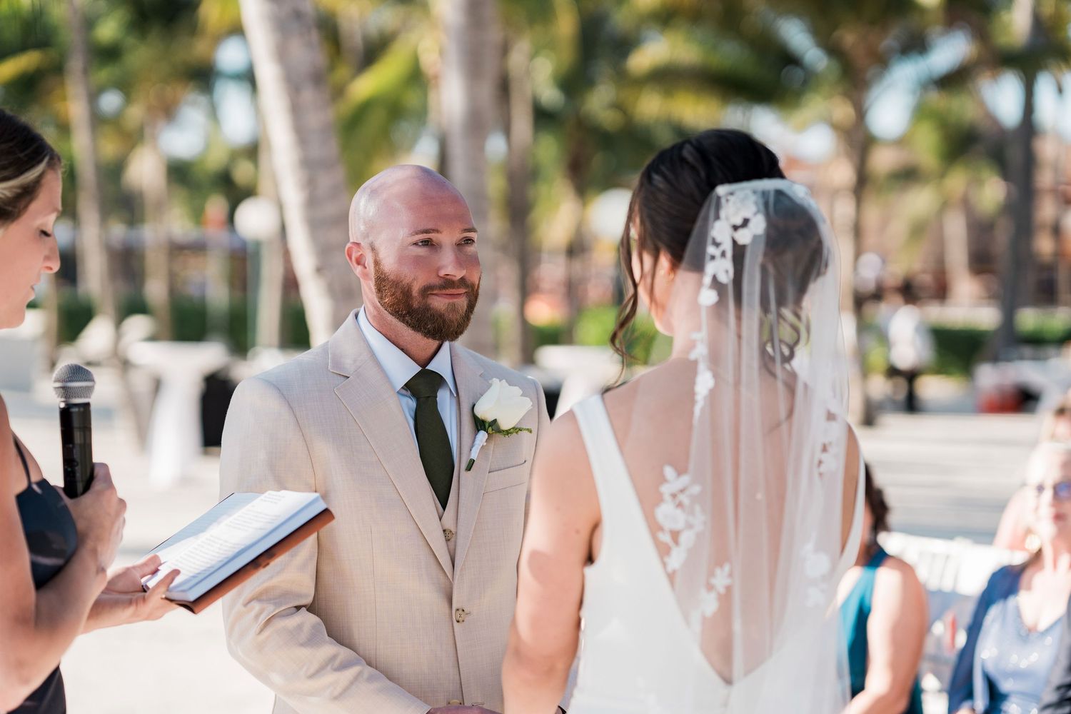 Groom in beige suit smiling at bride during outdoor beach wedding ceremony with palm trees.