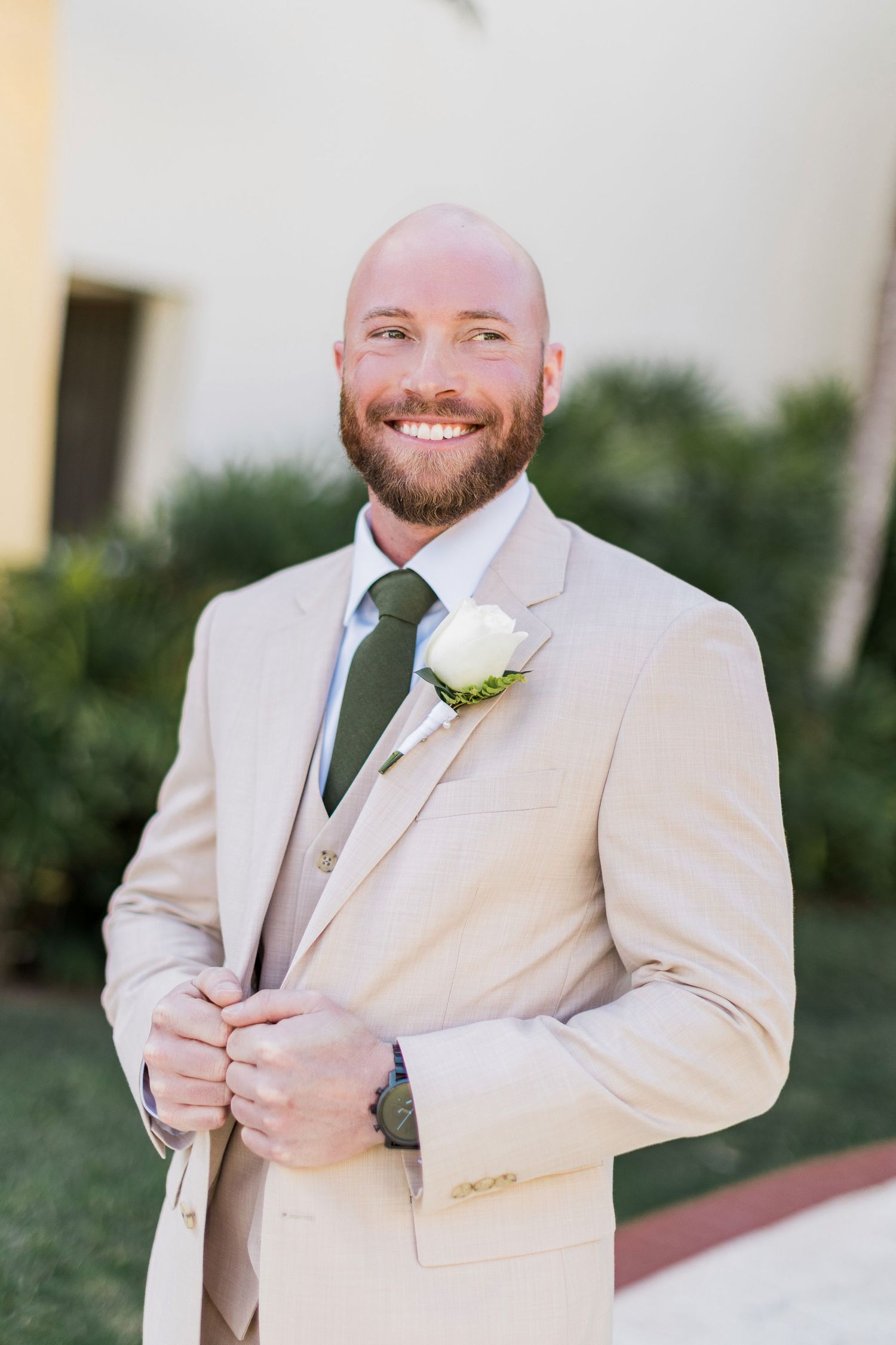 Smiling bald groom in beige suit with green tie and white rose boutonniere poses outdoors at wedding venue.