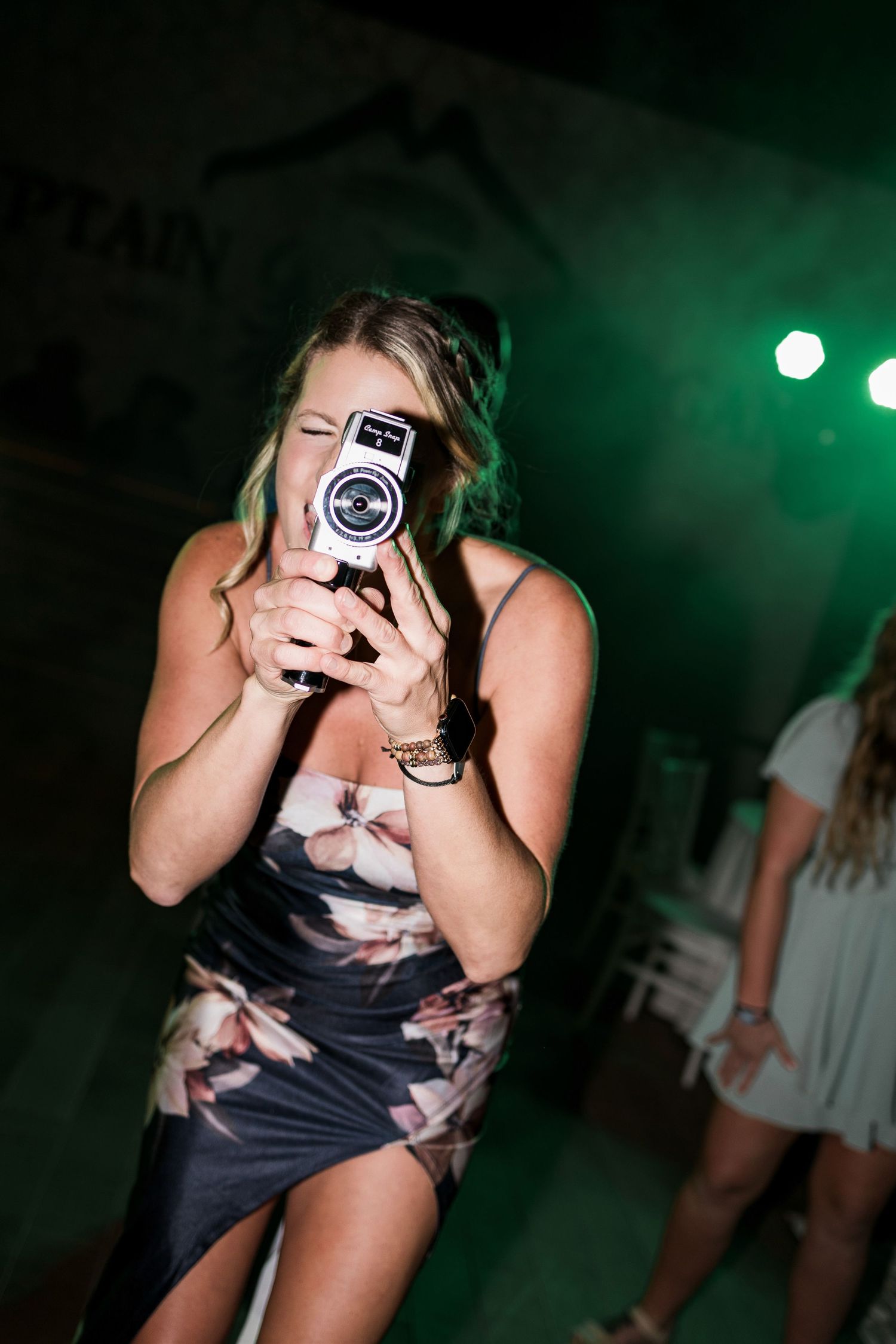 Woman in floral dress photographing with vintage camera at night party with green stage lighting.