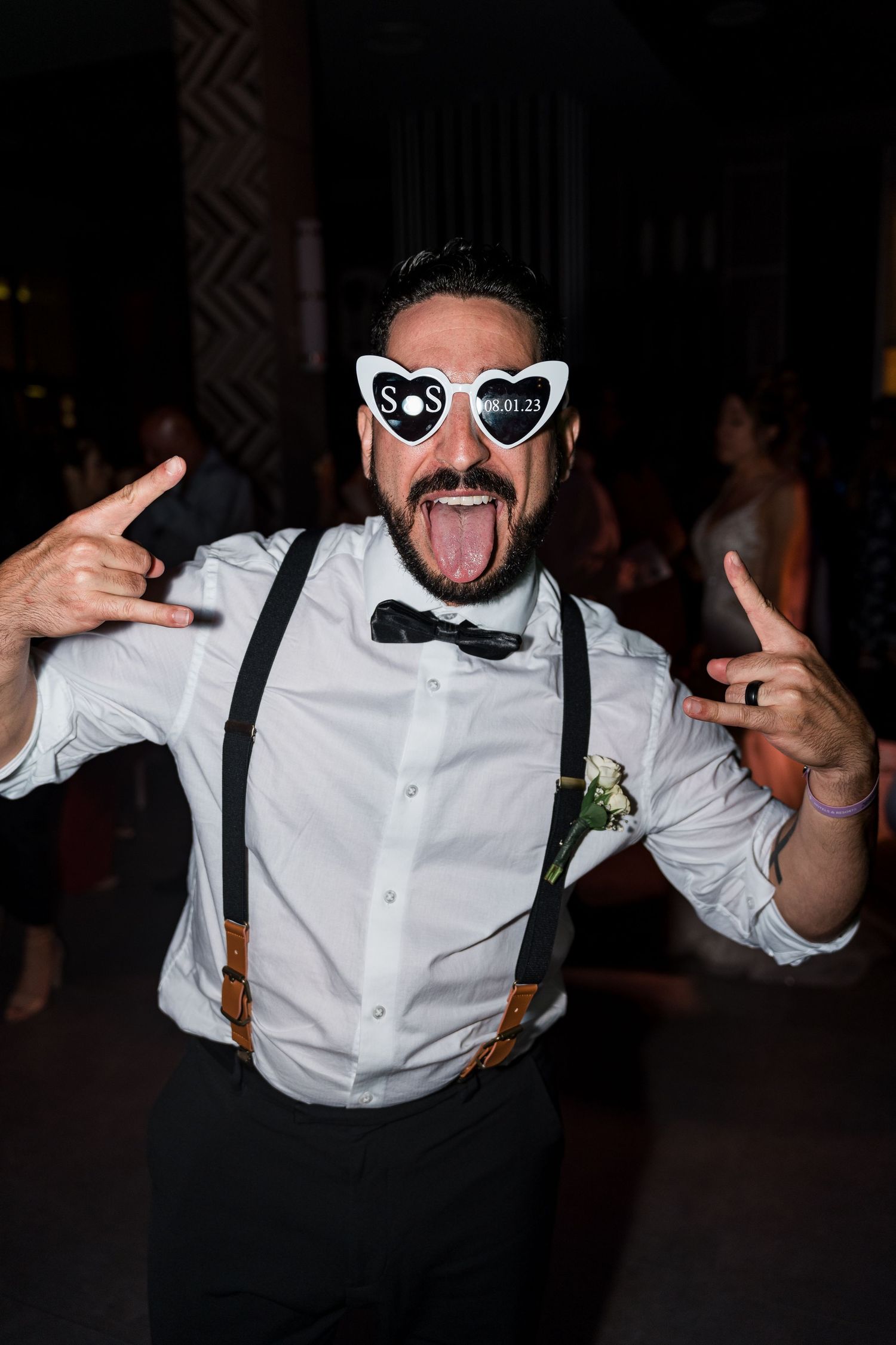 Wedding guest wearing heart-shaped S&S sunglasses and suspenders makes rock gesture at reception.