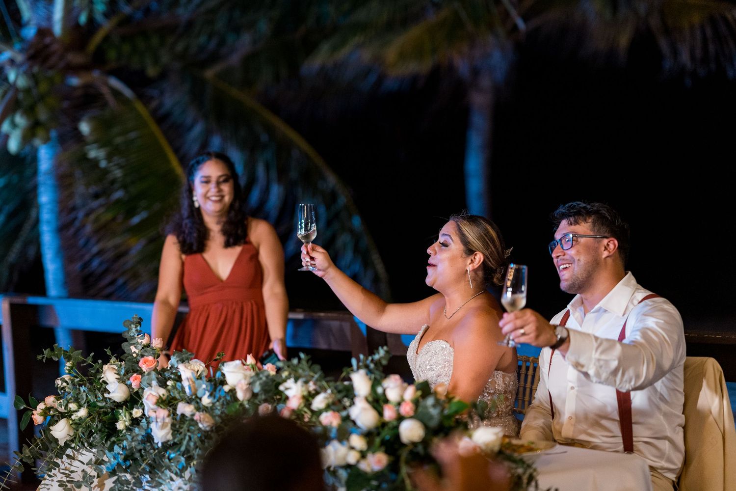 Bride and groom raising champagne glasses in toast with bridesmaid at tropical beach wedding reception.