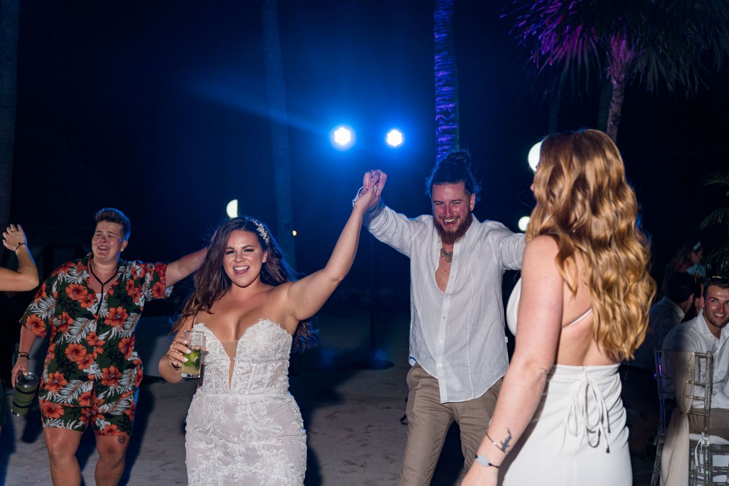 Bride in white gown dances joyfully with guests at an outdoor nighttime beach wedding reception.