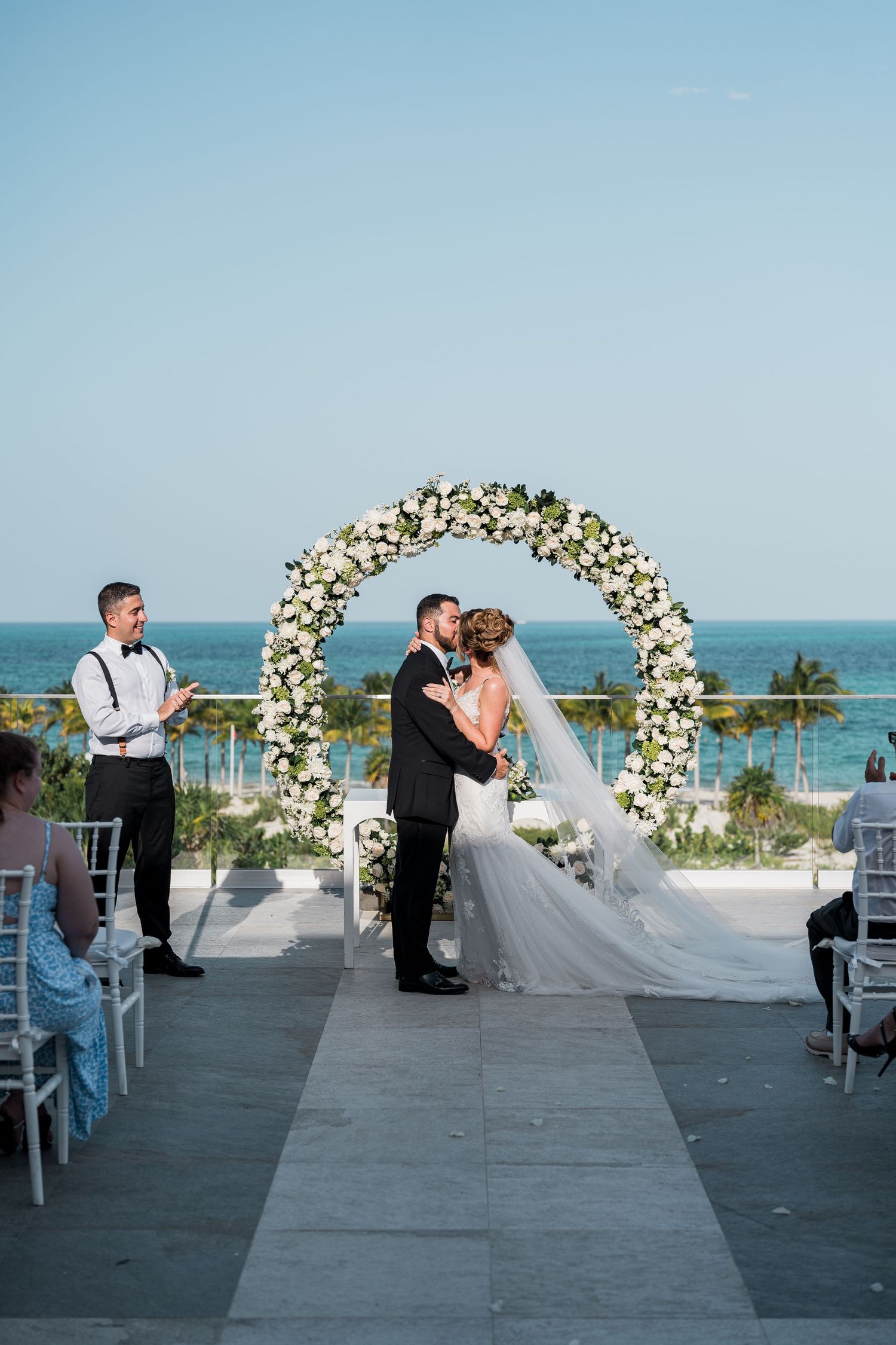 Bride and groom embrace during first kiss under circular white rose arch by the ocean.