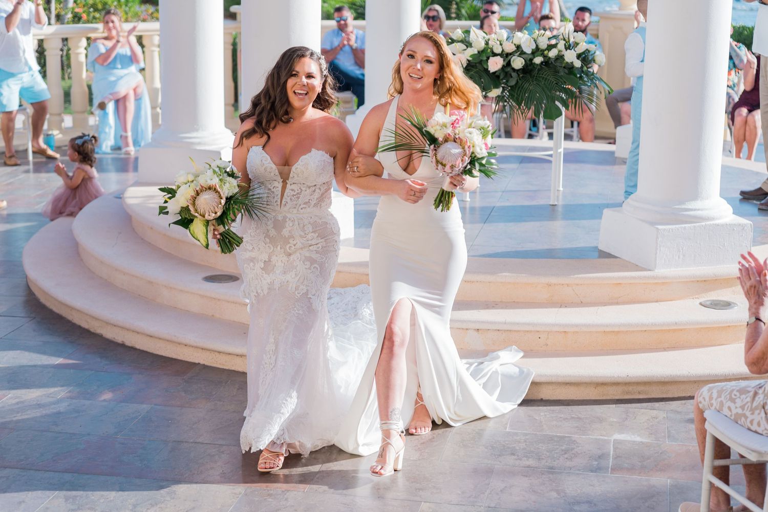 Two brides smiling and walking with bouquets after their tropical beachside wedding ceremony.