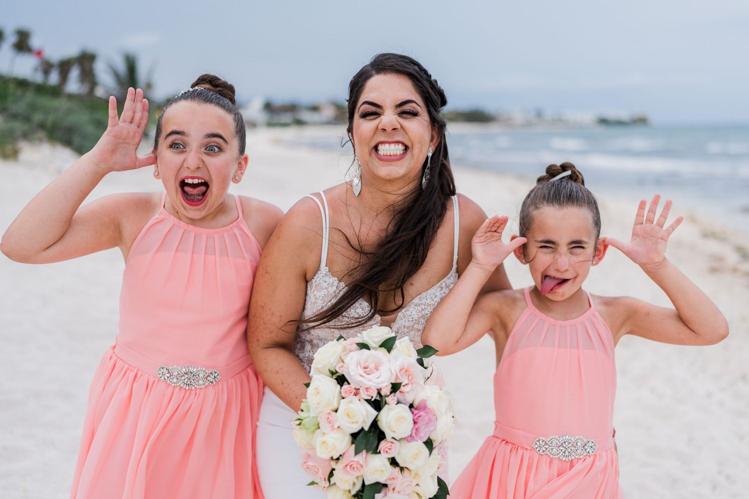 Bride holding white bouquet laughing with two young flower girls in coral pink dresses on a sunny beach.