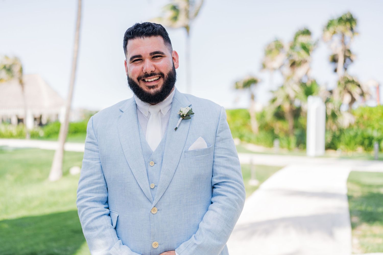 Smiling groom in light blue suit with boutonniere poses outdoors at tropical beach wedding venue.