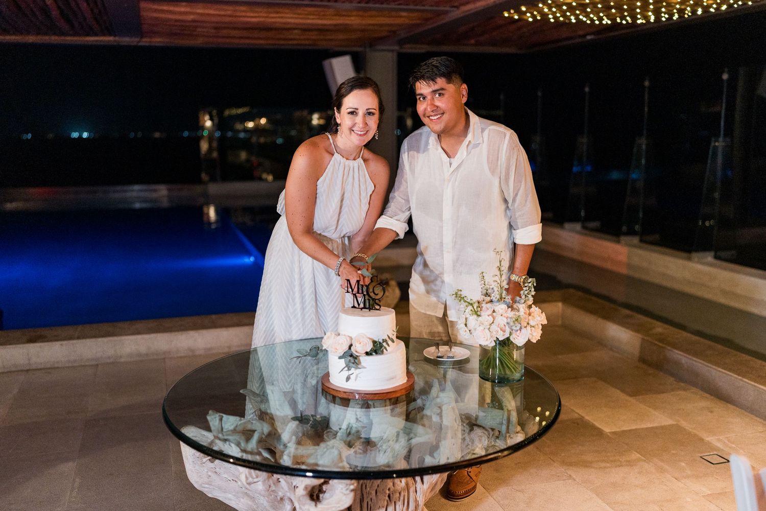 Bride and groom smile together while cutting their white wedding cake at an outdoor poolside reception.