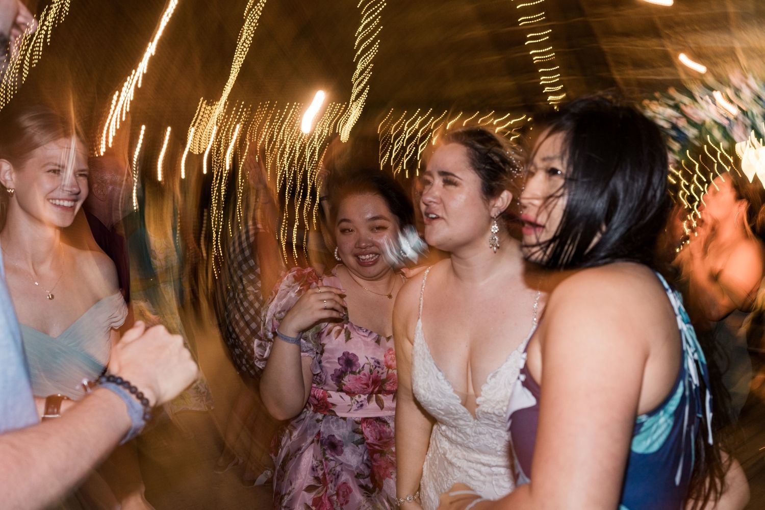 Wedding guests laugh and dance together under spinning string lights at lively reception.