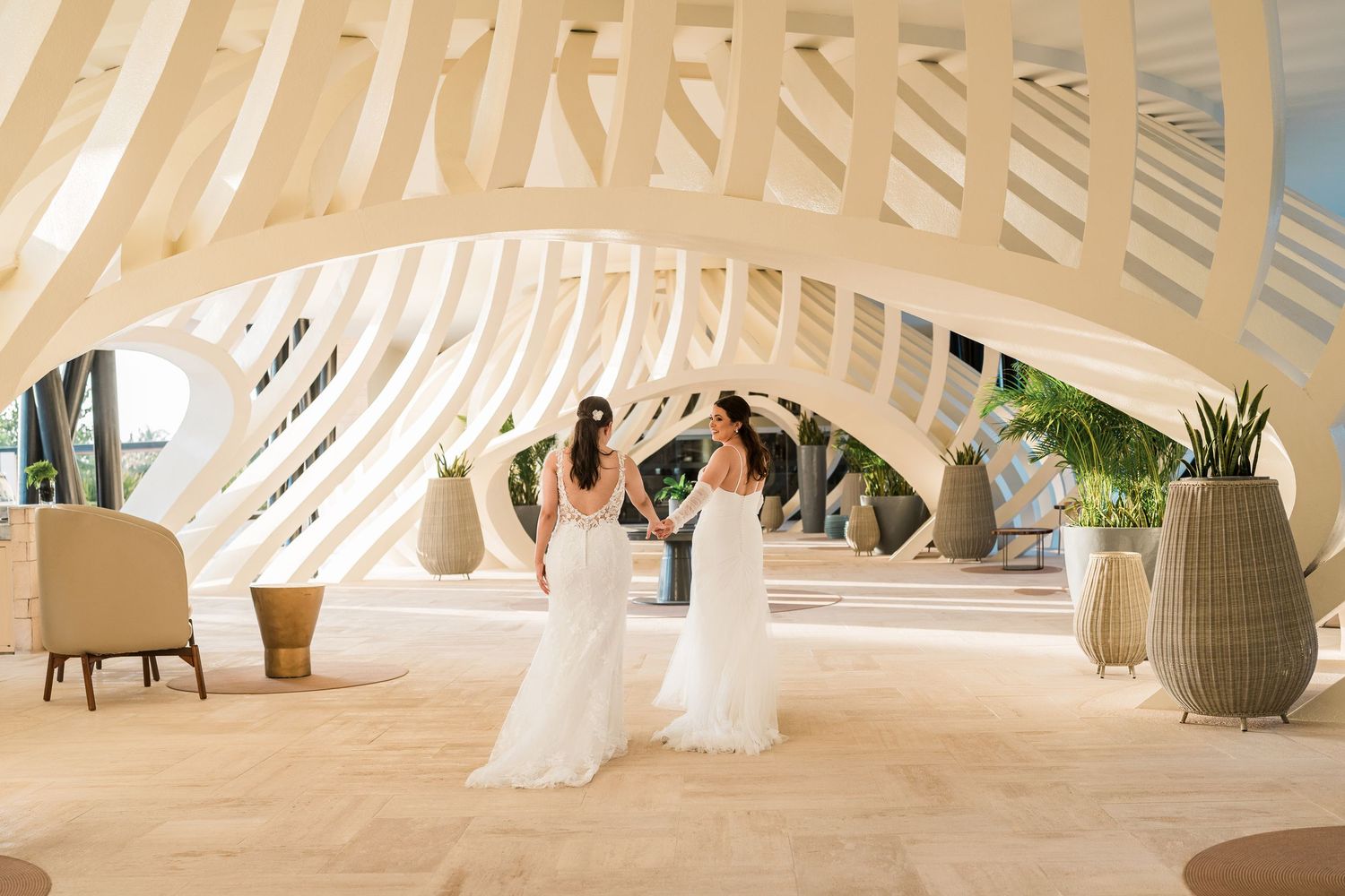 Two brides in white gowns hold hands under an elegant curved white archway at a luxury wedding venue.