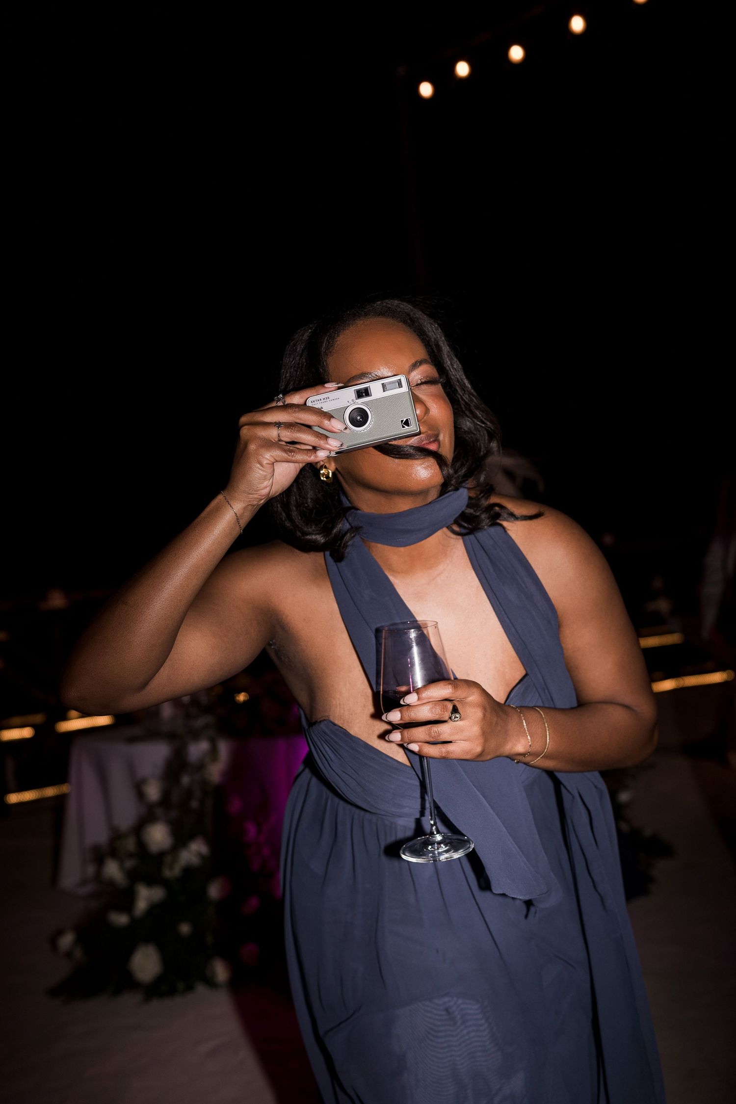 Woman in blue halter dress holding wine glass and film camera at evening wedding reception.