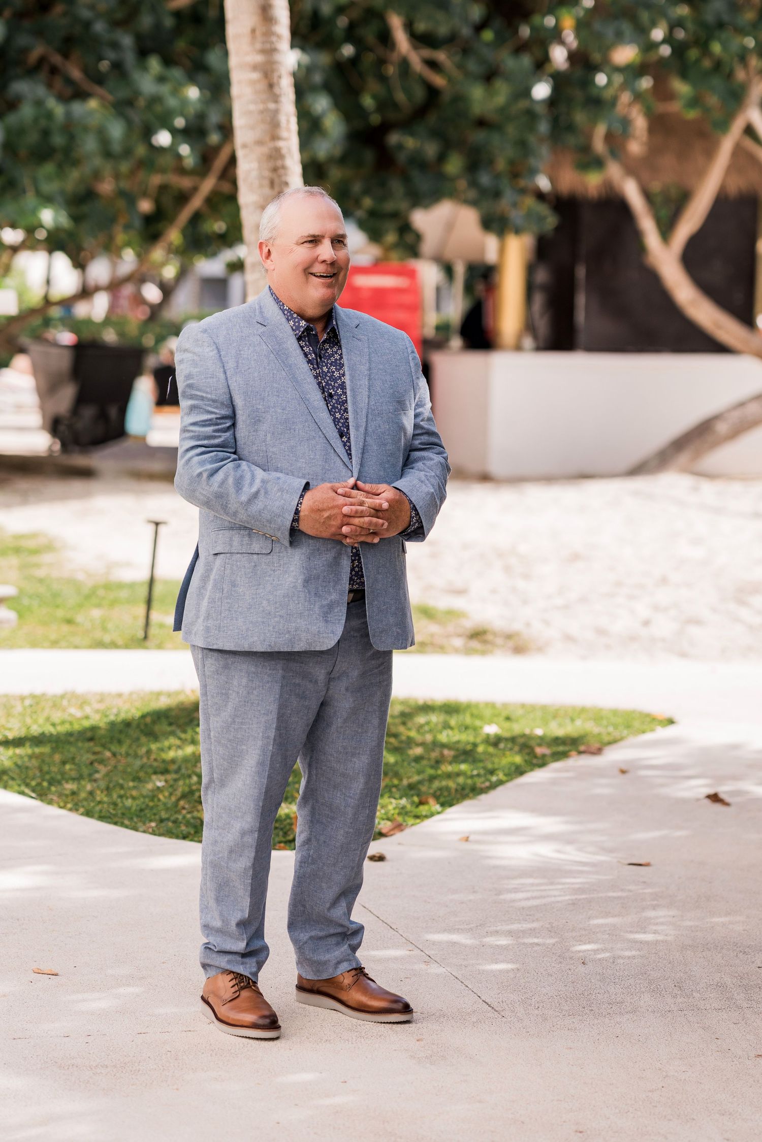 Smiling groom in light blue linen suit with floral shirt standing outdoors on a sunny wedding day.