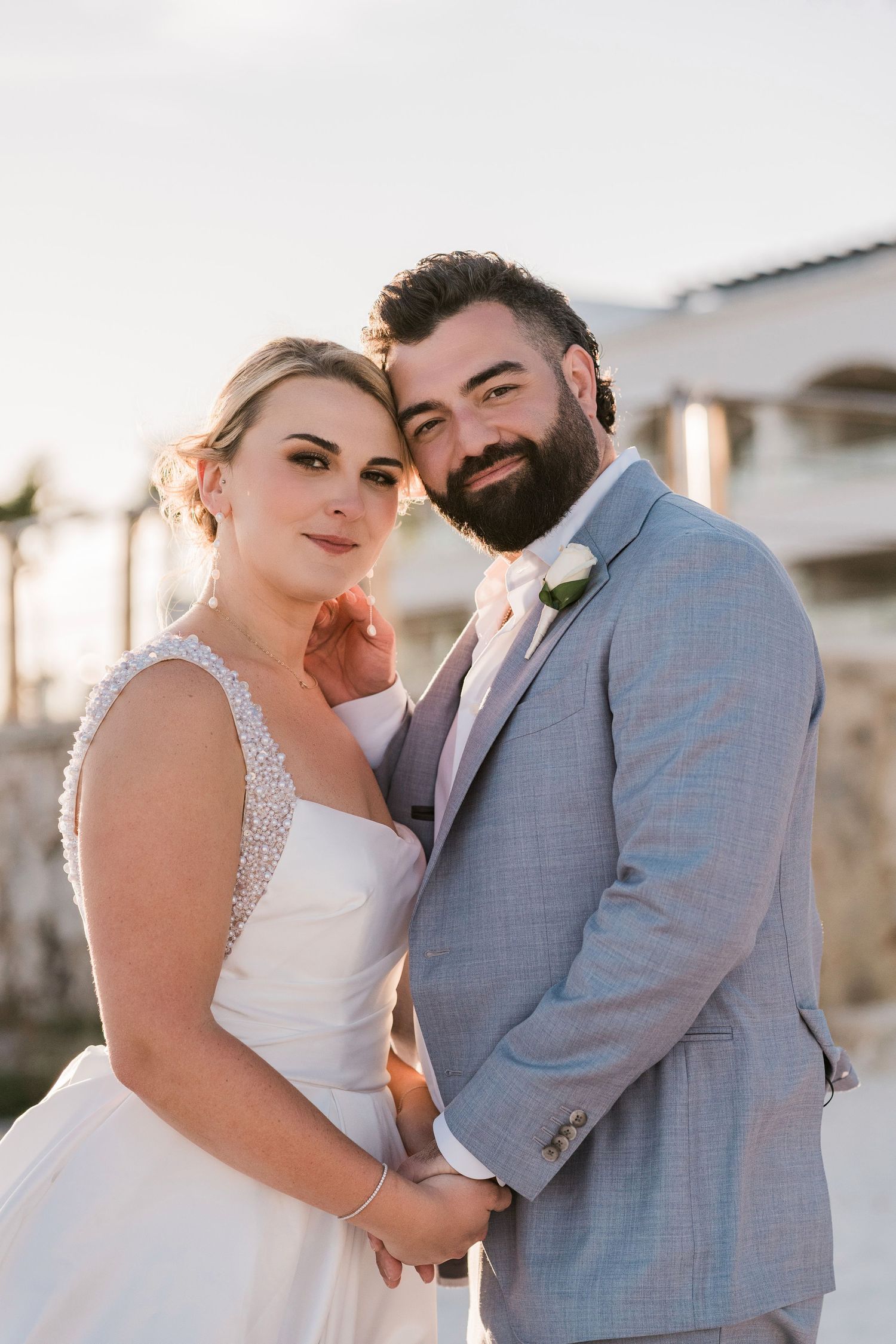 A bride and groom pose together outdoors, she in a beaded white gown and he in a light gray suit with a white boutonniere.