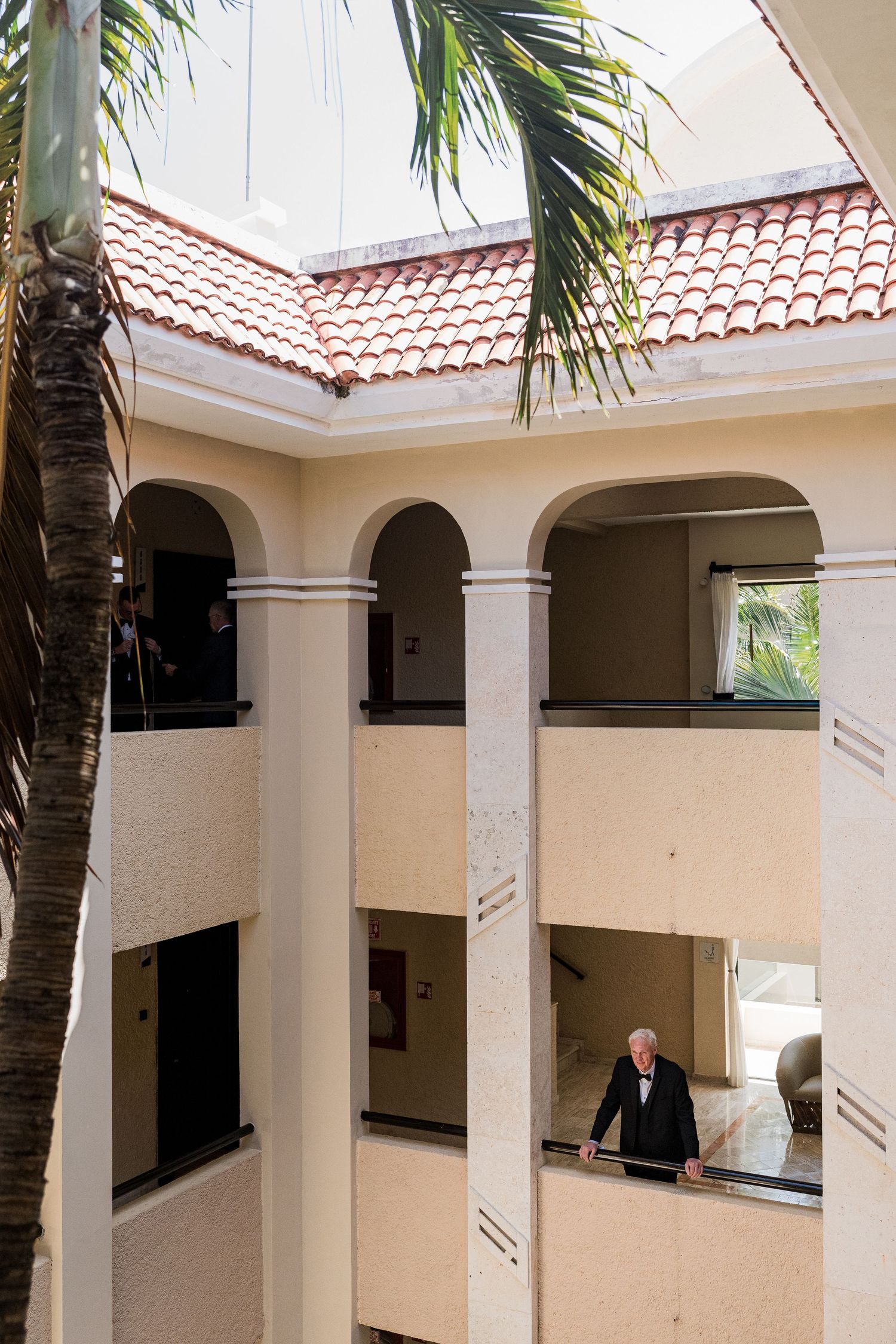 Elegant Mediterranean-style hotel atrium with arched columns, red tile roof, palm trees, and a senior man standing on the balcony.