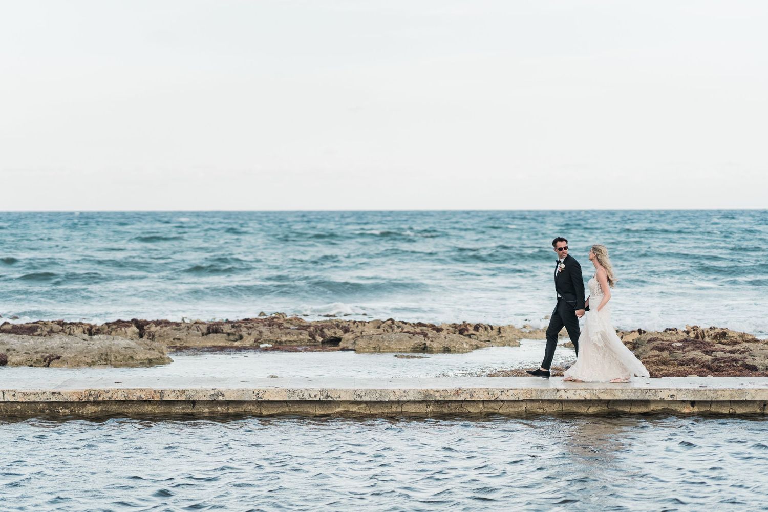 Bride and groom walking on a seaside pier with ocean waves in the background on their wedding day.