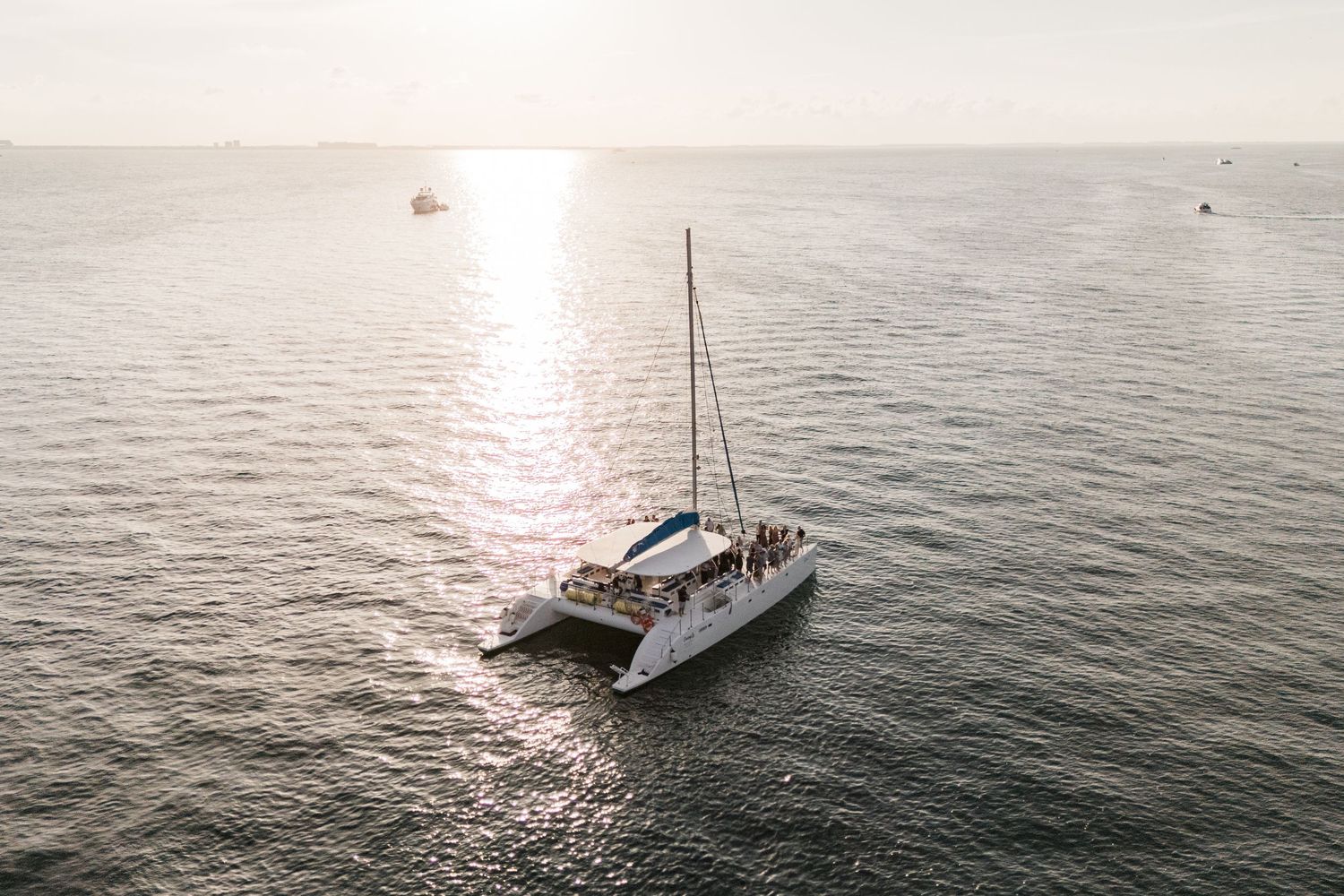 Aerial view of a catamaran sailboat with passengers cruising calm ocean waters at golden sunset.