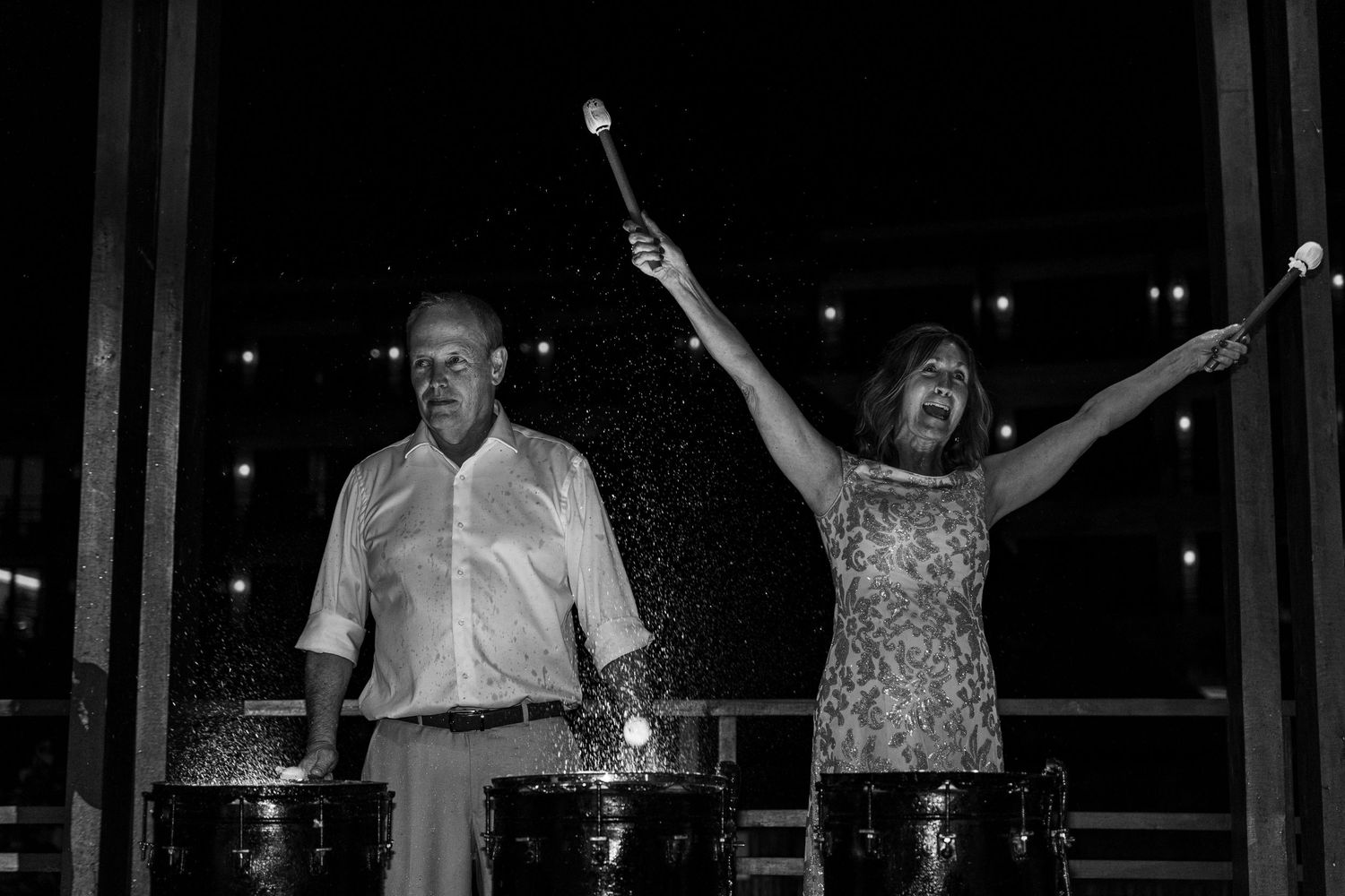 Black and white photo of a man and woman enthusiastically playing steel drums at a nighttime outdoor event.