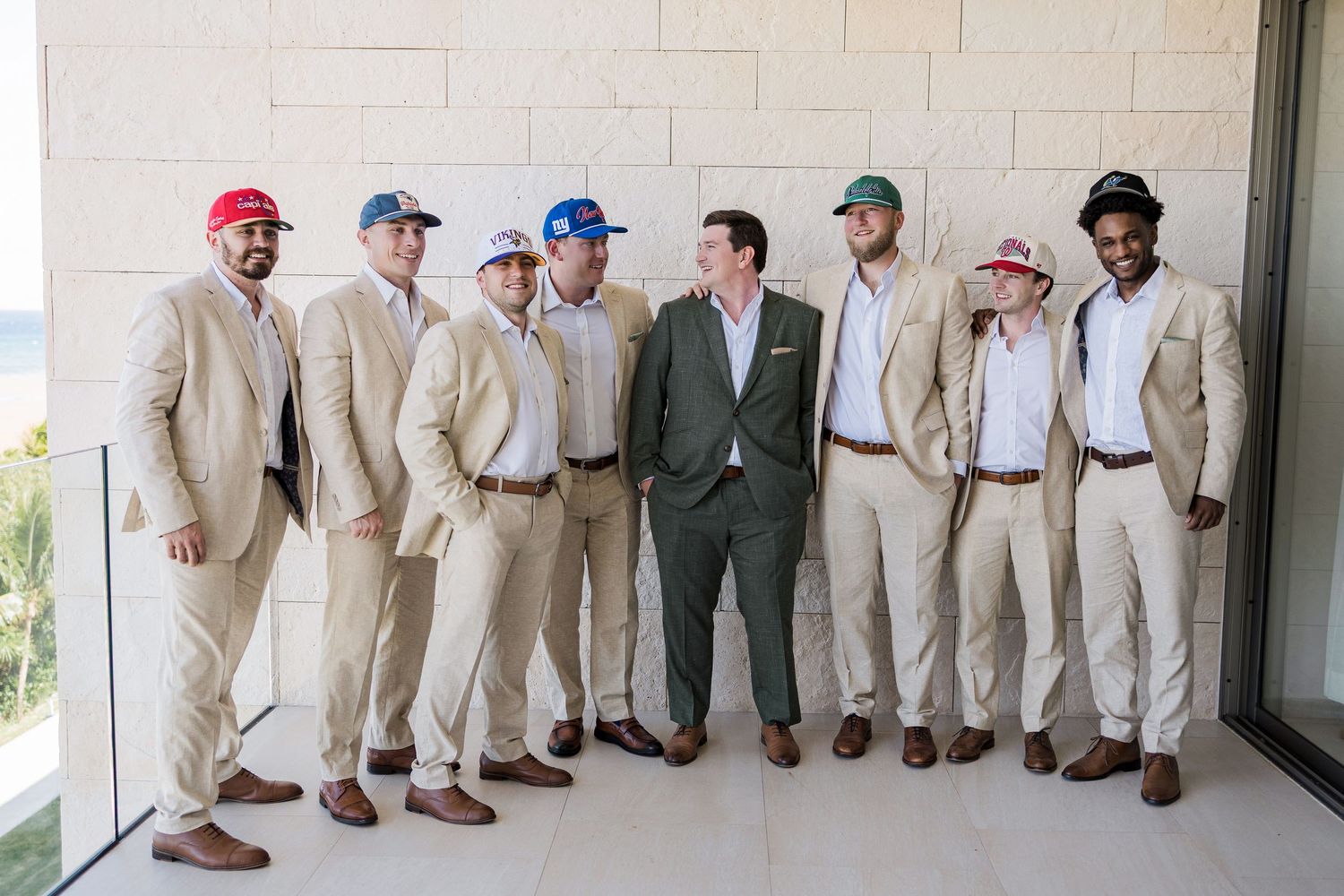 Groomsmen in tan suits and colorful baseball caps pose with groom in green suit outdoors at wedding venue.