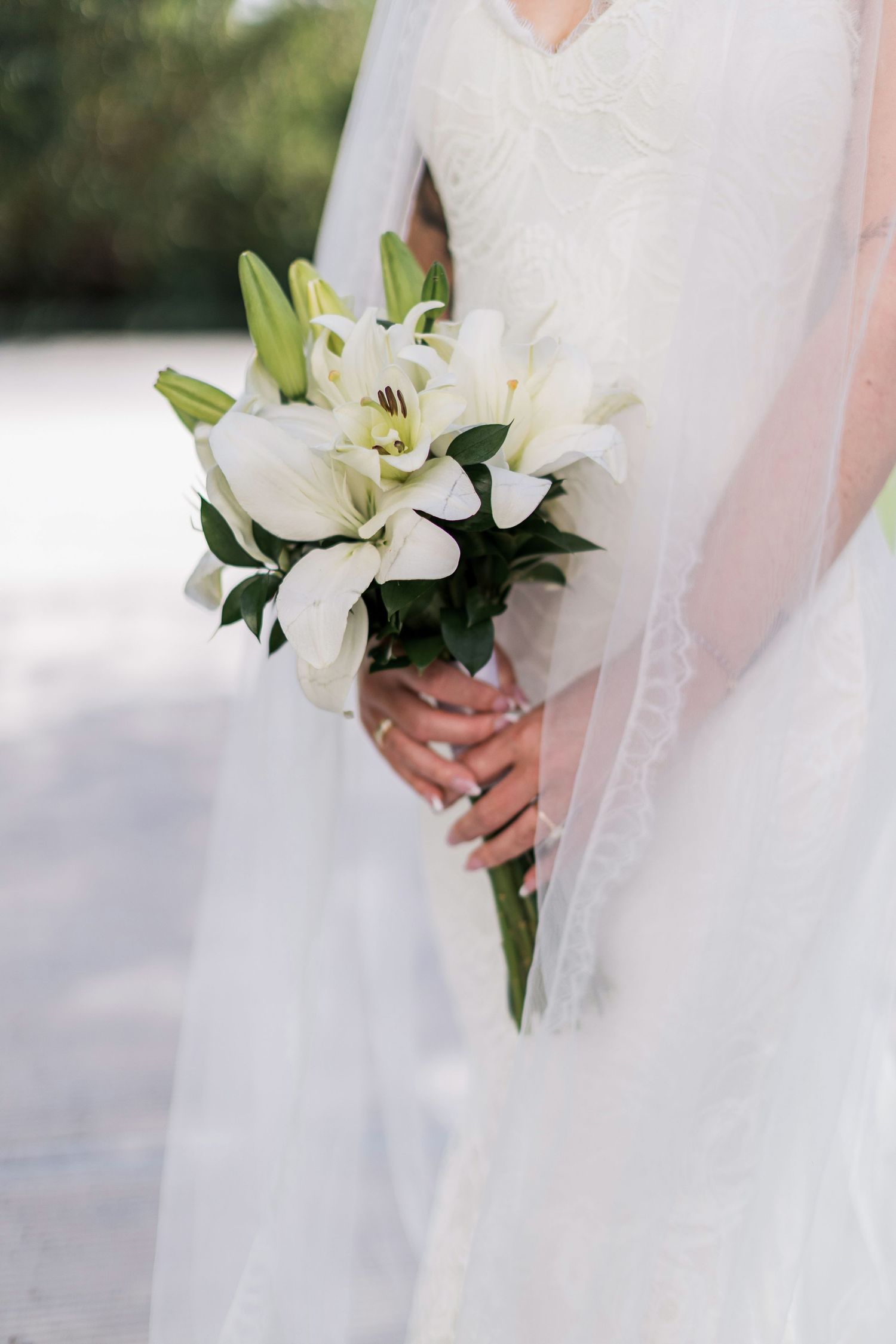 Bride in white lace gown holding elegant bouquet of white lilies with green leaves on her wedding day.