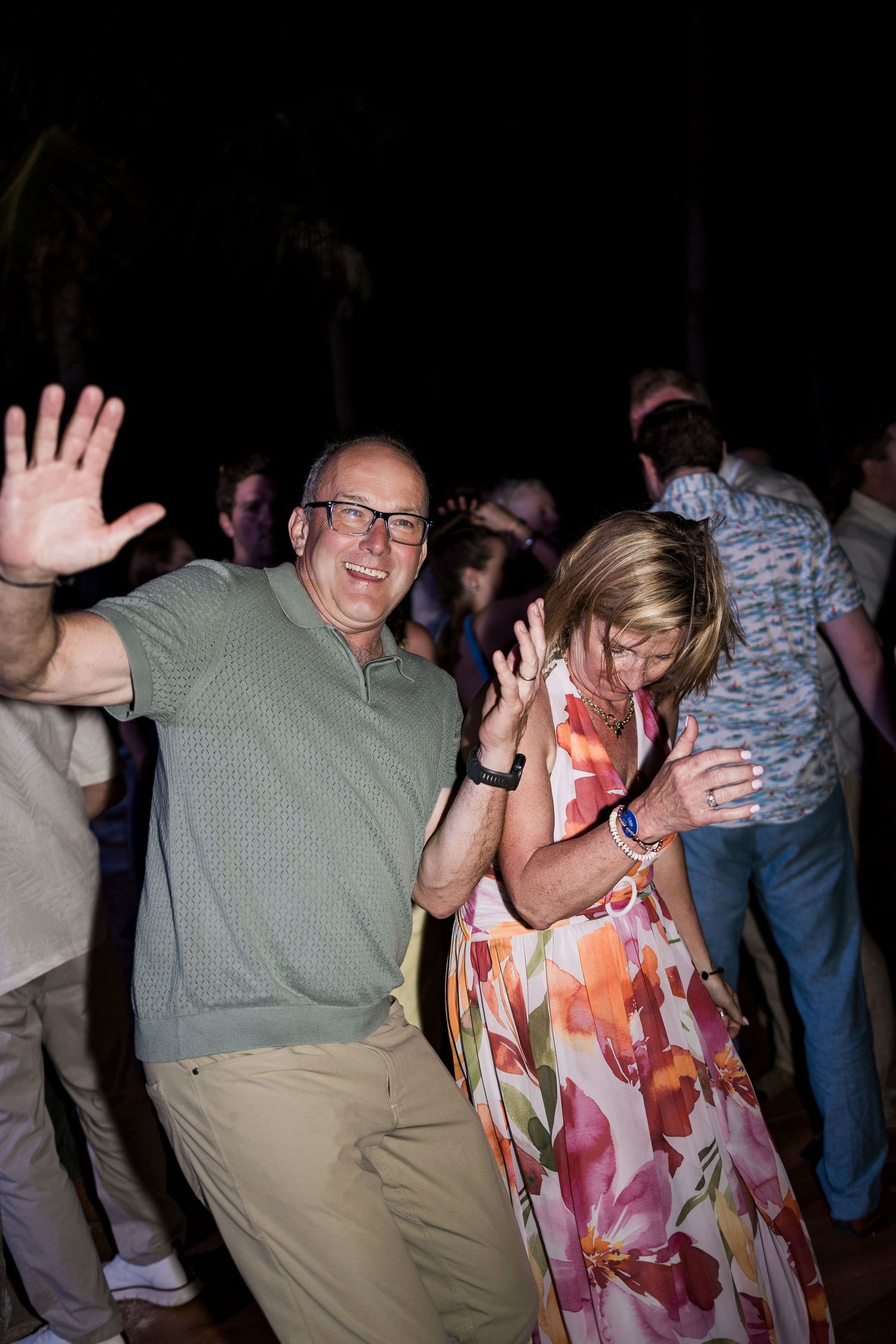 Couple dancing joyfully at an outdoor evening party, man in green polo raising hands, woman in floral dress.