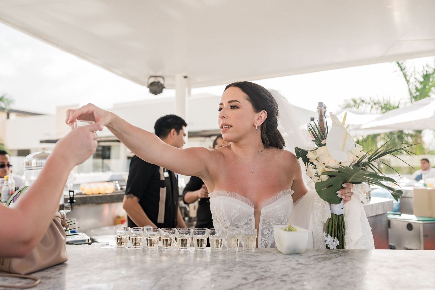 Bride in strapless gown arranging shot glasses on marble bar with white floral arrangement at outdoor wedding reception.