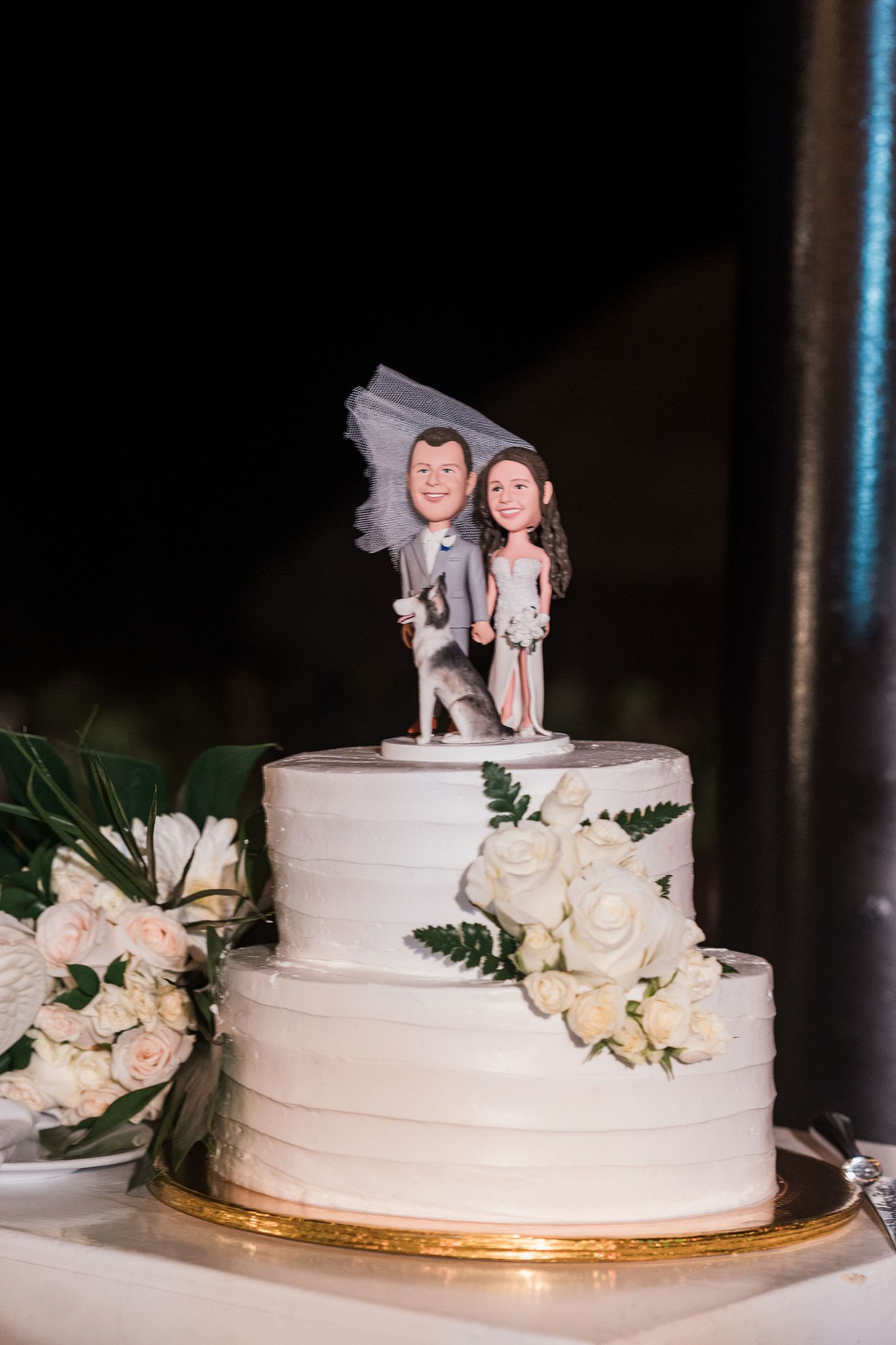 Two-tier white wedding cake with custom bobblehead cake toppers of bride and groom with dog, adorned with white roses and ferns.