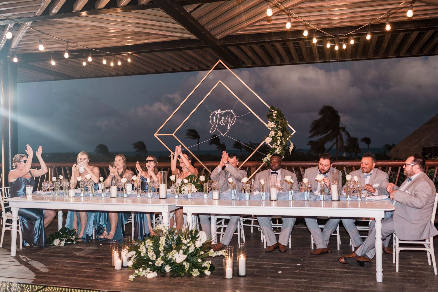 Elegant outdoor wedding reception with bridal party seated at long white table under string lights with geometric backdrop.