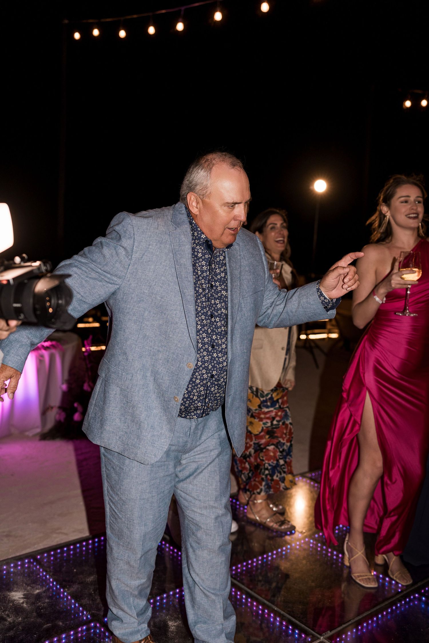 Elderly man in gray suit dancing at elegant evening reception with guests in formal attire on illuminated dance floor.