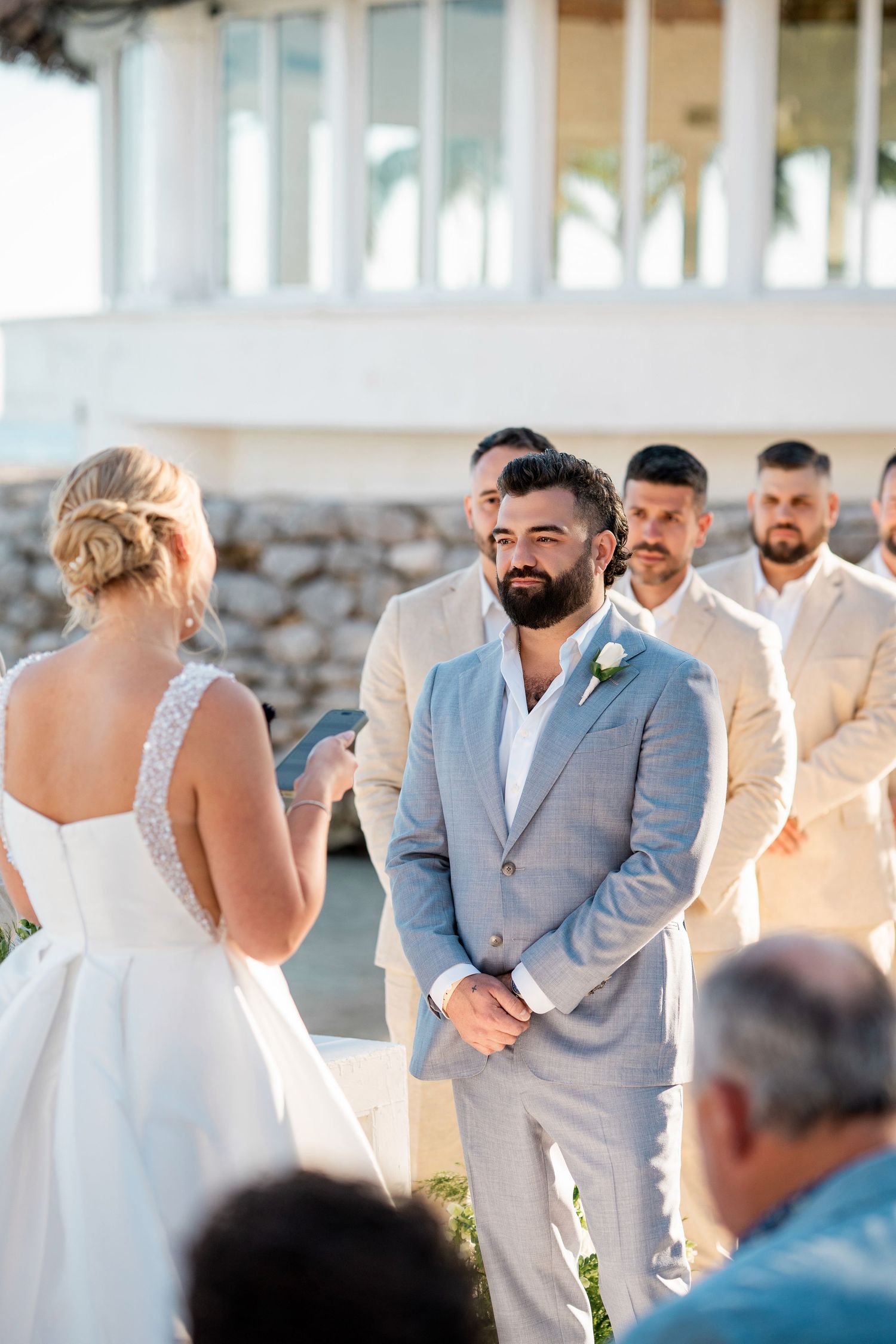 Groom in gray suit smiling at bride during outdoor wedding ceremony with groomsmen in beige suits in background.