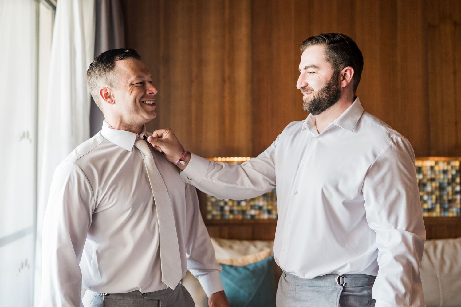 Two men in white dress shirts smile as one helps the other adjust his tie while getting ready for a wedding.