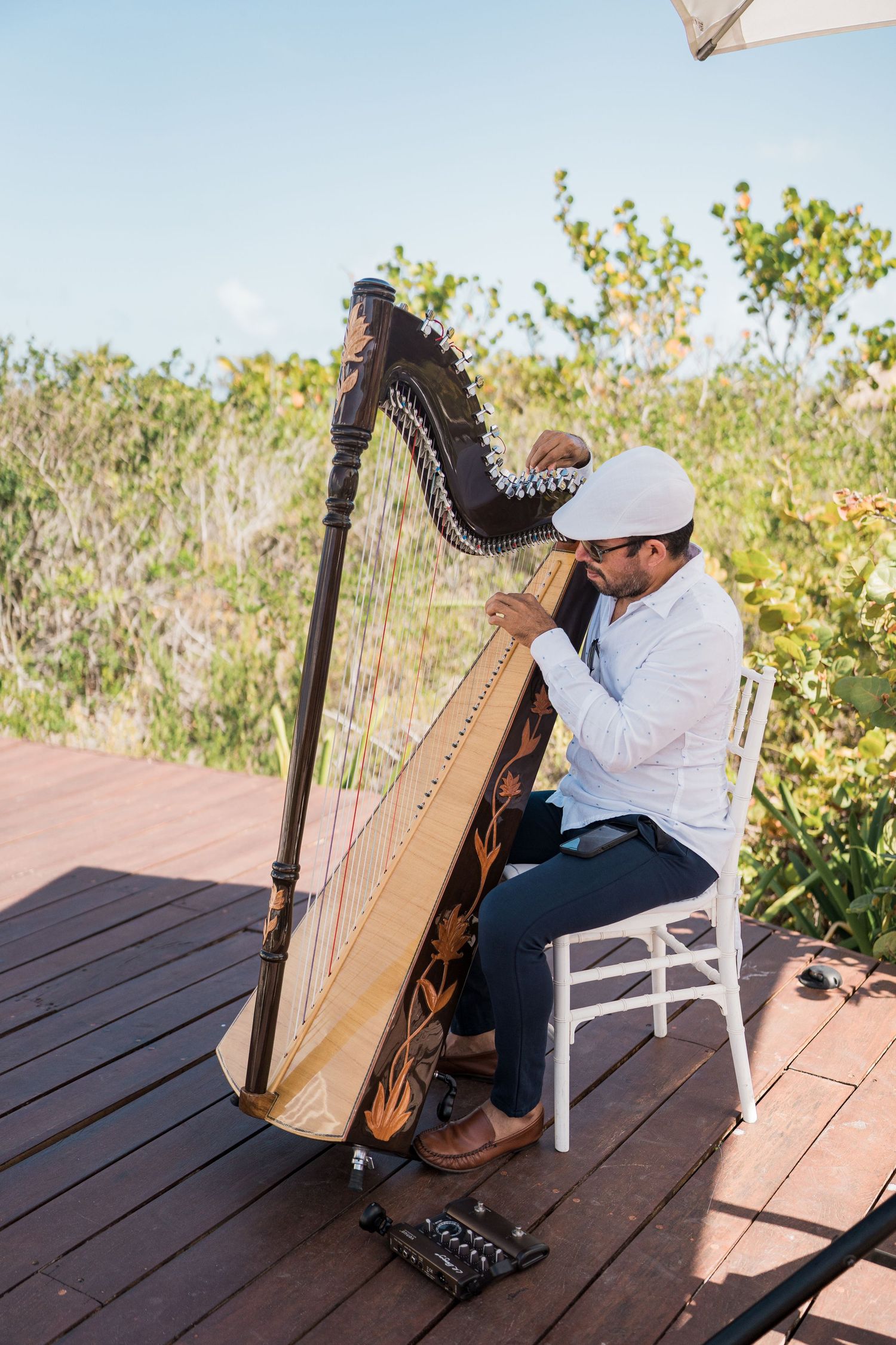 Man in white shirt and cap playing a decorated harp on a wooden deck outdoors with lush greenery behind him.