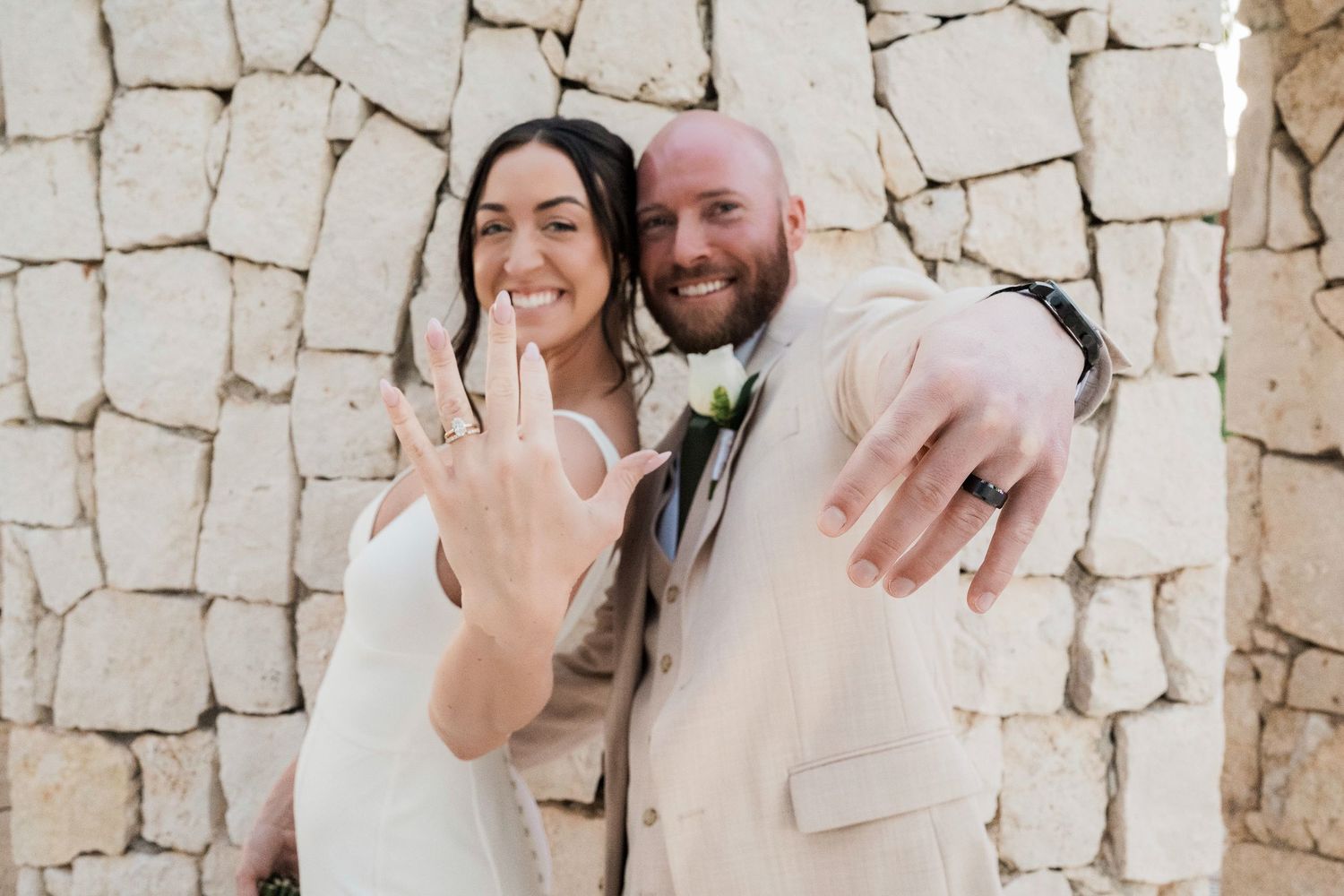 Newlywed couple smiling and showing off wedding rings against a stone wall backdrop, barceló Maya palace wedding