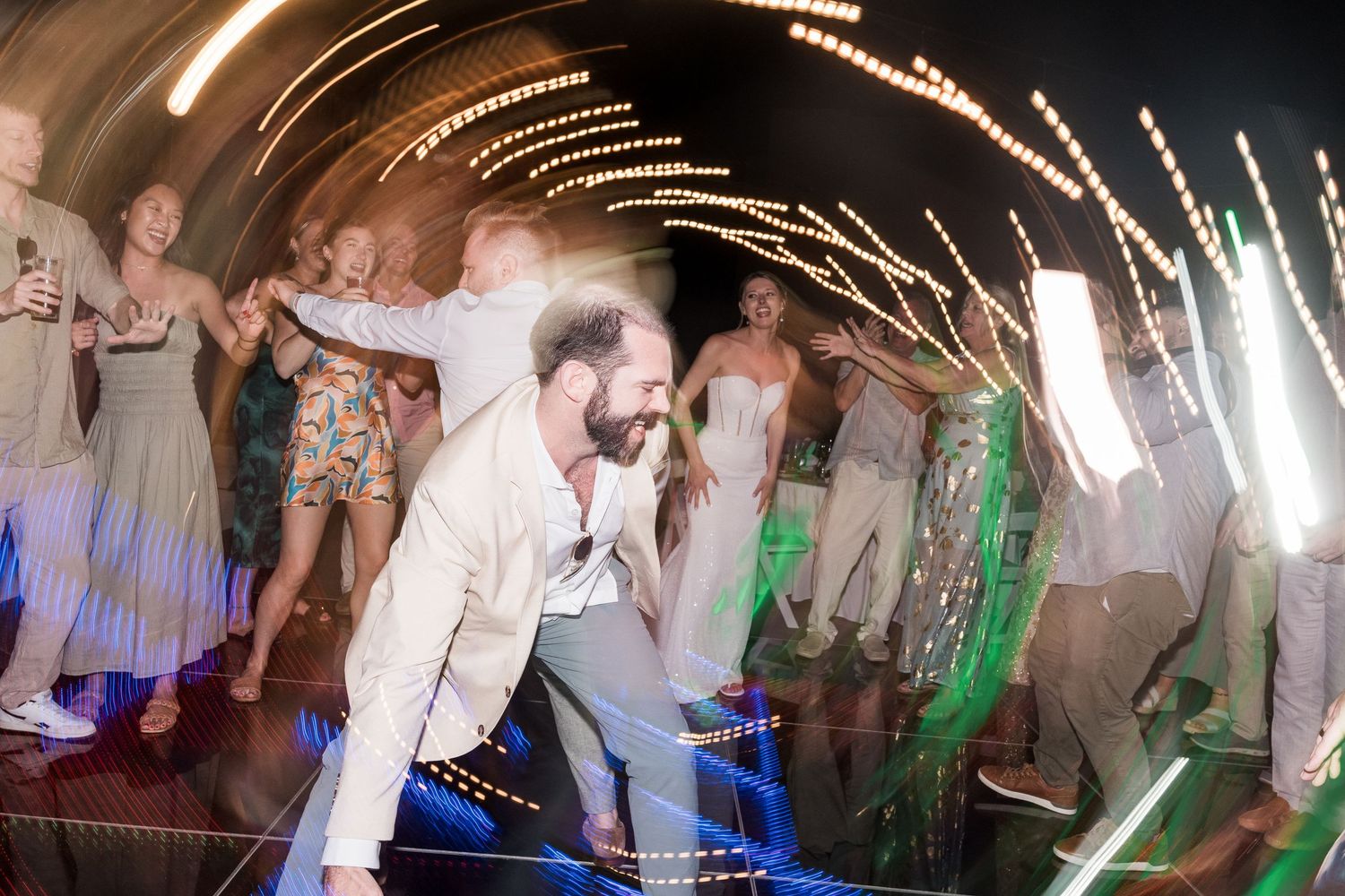 Energetic wedding reception dance floor with bride and groom celebrating with guests under swirling light trails.