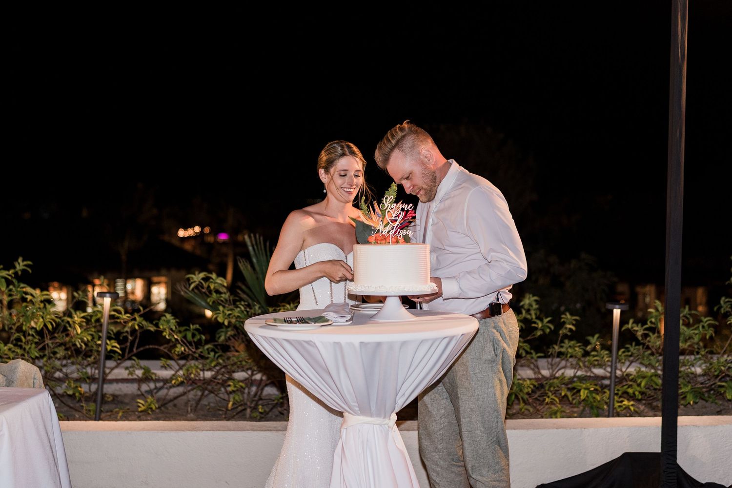 Bride and groom cutting their white wedding cake together at an elegant outdoor nighttime reception.