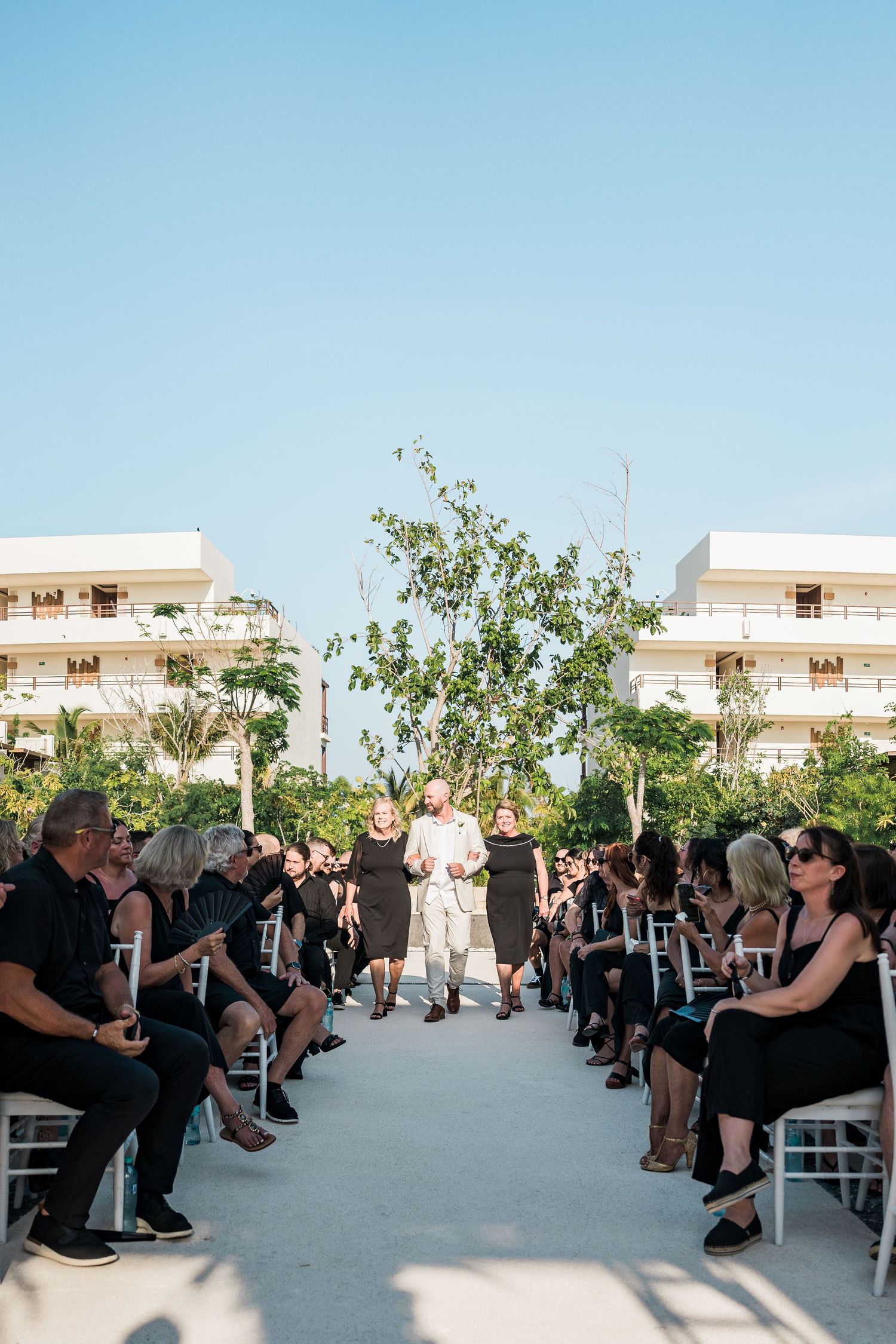 Outdoor wedding ceremony with guests in black attire seated along a white aisle, flanked by trees and resort buildings.