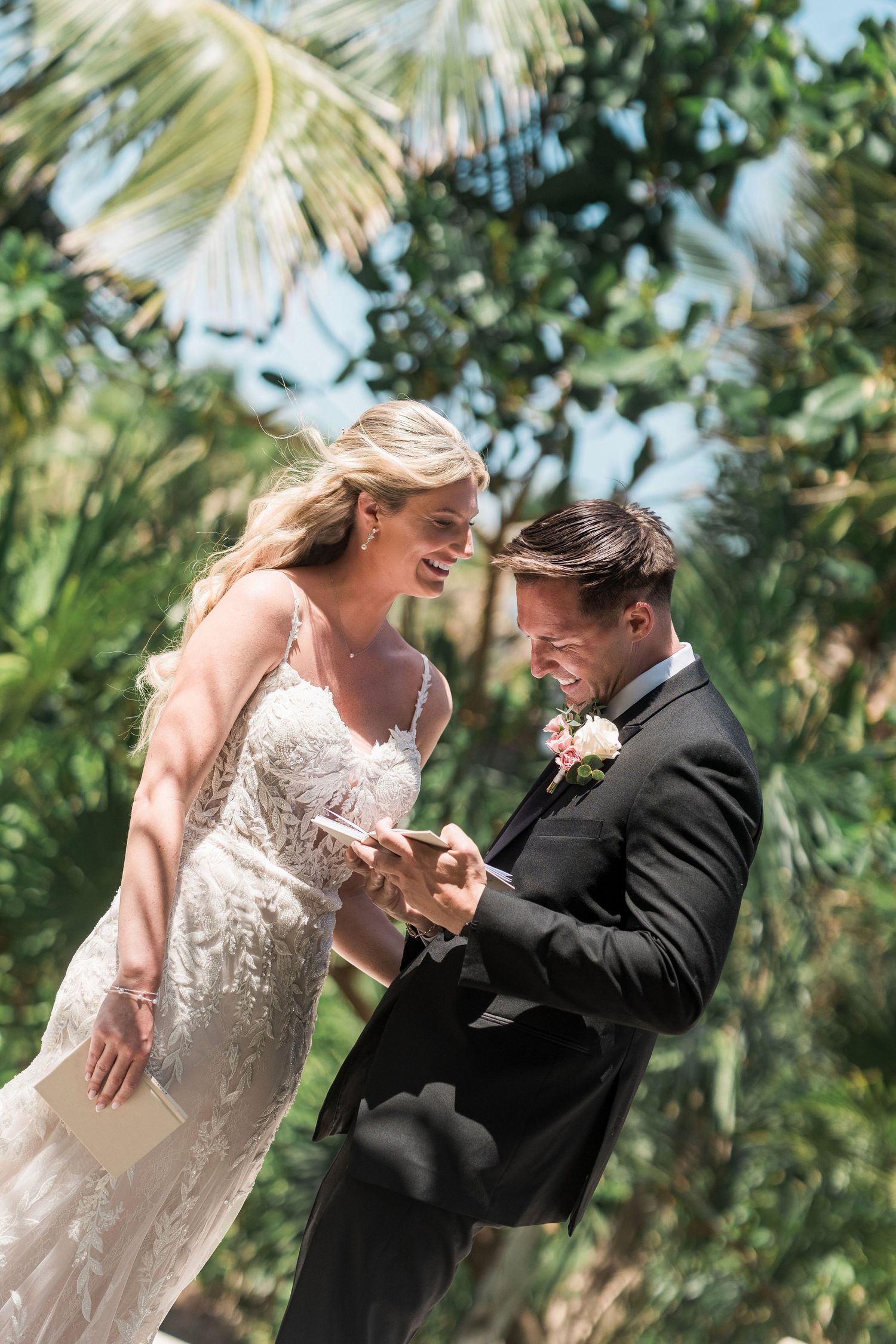 Bride and groom exchange rings outdoors at tropical wedding ceremony, smiling joyfully with palm trees in background.