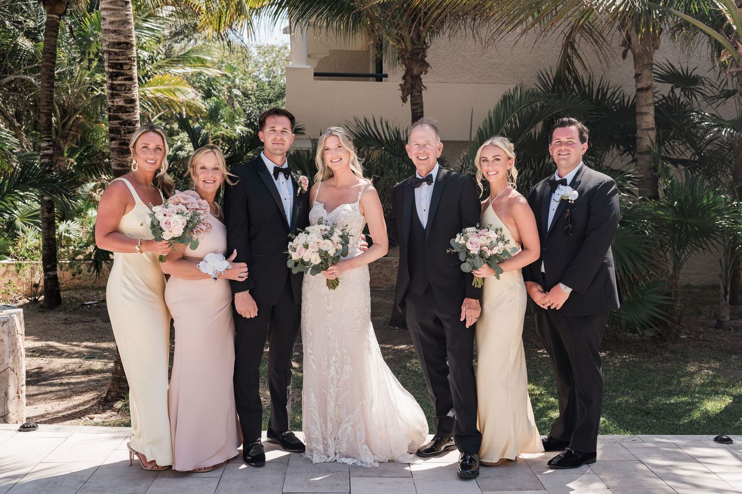 Wedding party posing outdoors with bride and groom, bridesmaids in champagne gowns and groomsmen in black tuxedos holding white bouquets.