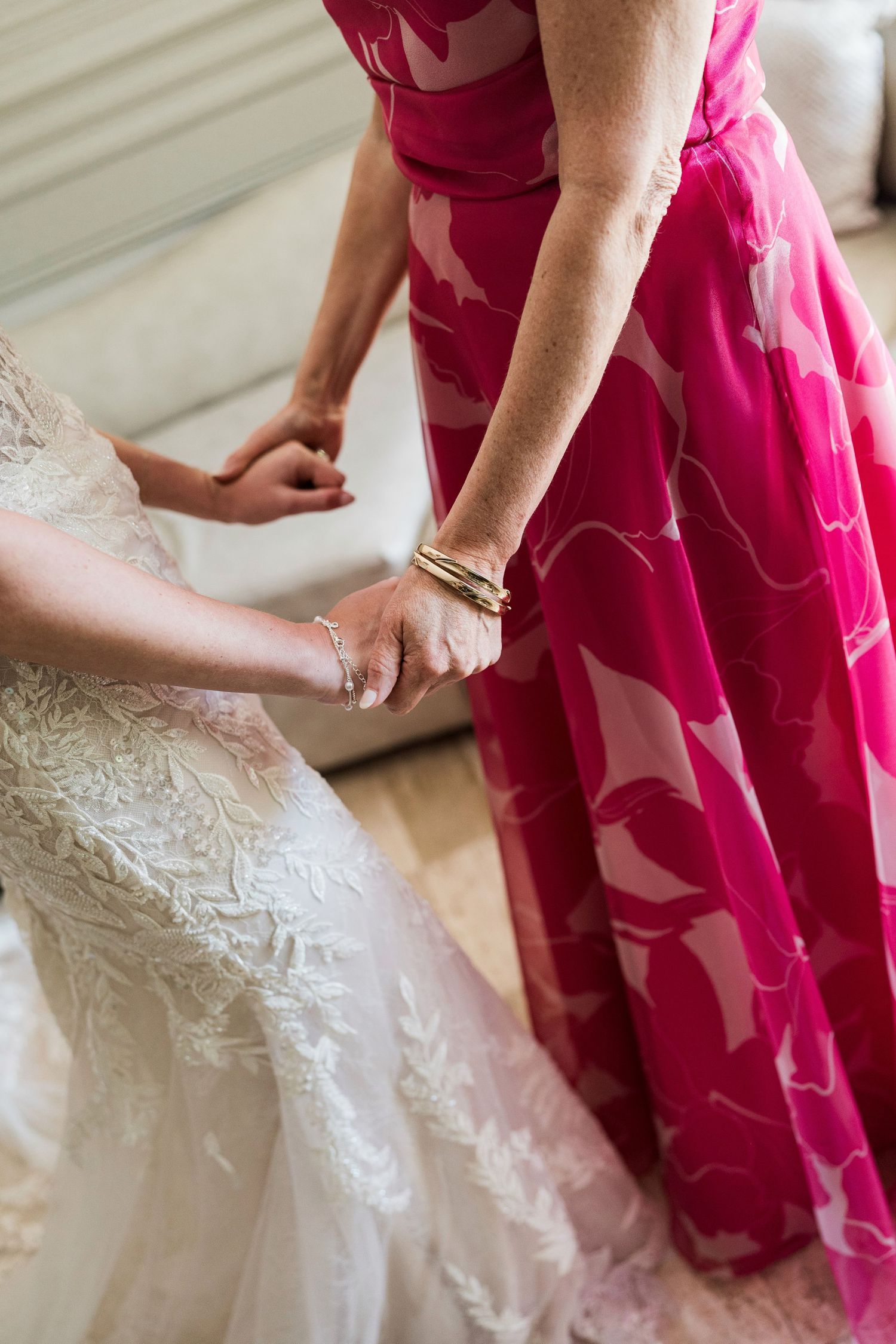 Bride in lace wedding gown holds hands with woman in pink floral dress during wedding preparation.