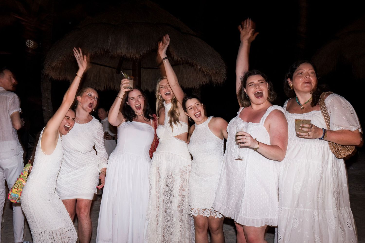 Group of women in white dresses celebrating at an outdoor night party, raising hands and holding drinks.