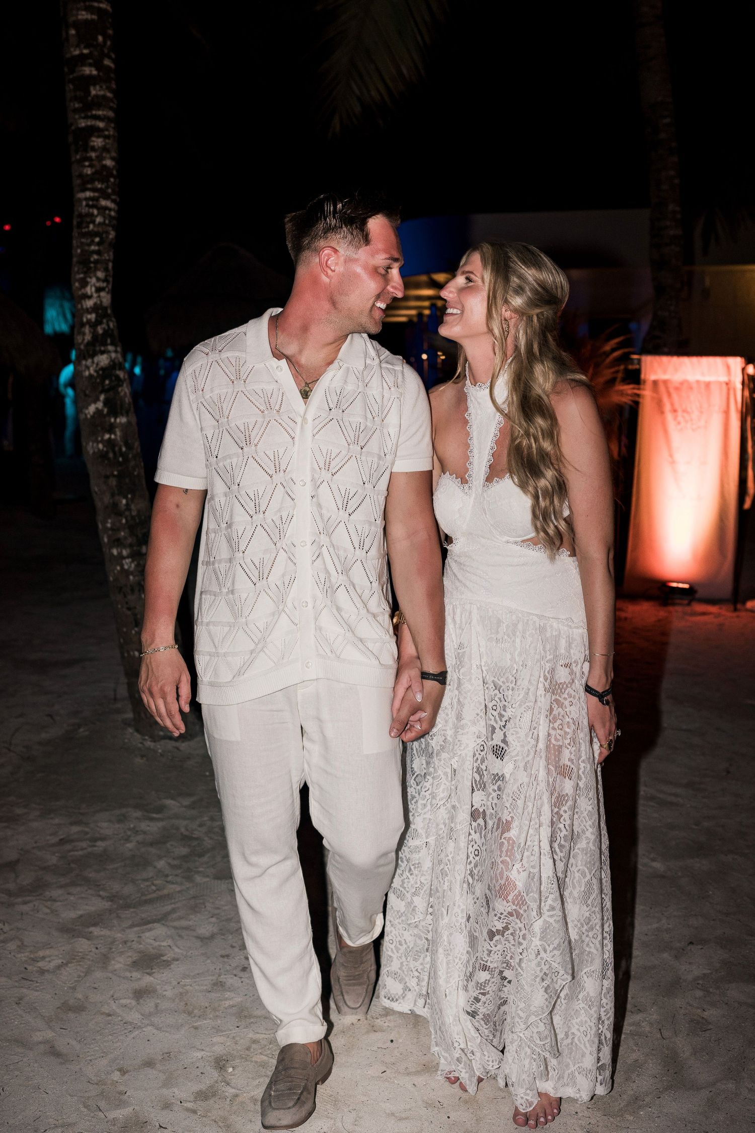 Couple in matching white outfits holding hands and smiling at each other on a sandy beach at night.