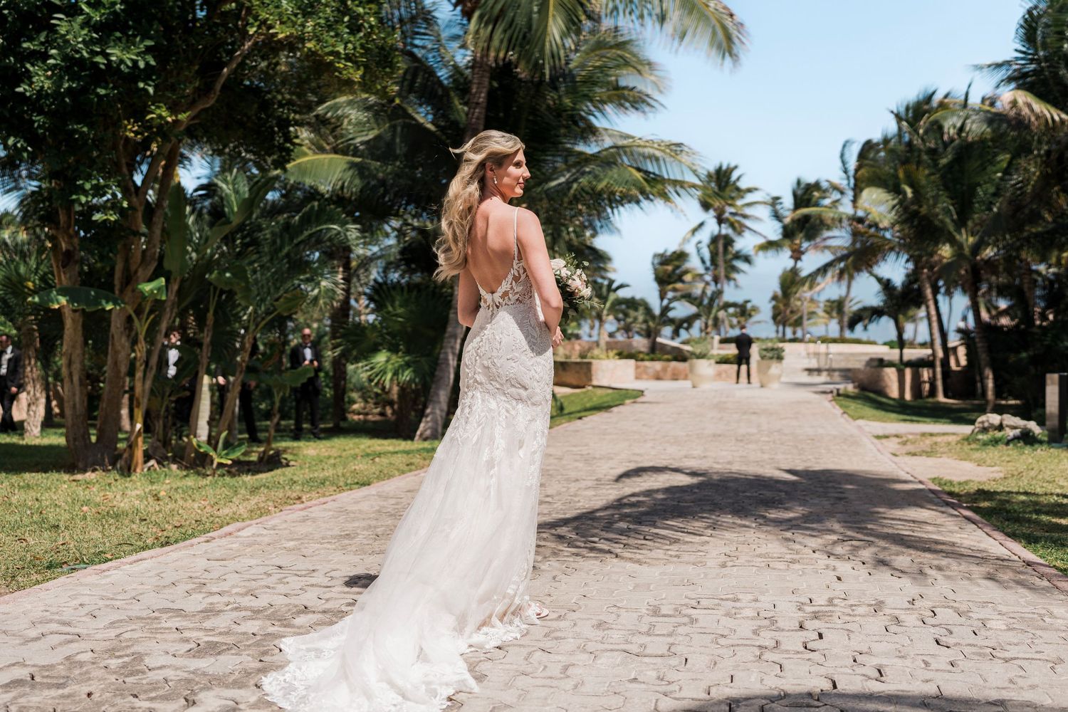 Bride in backless lace wedding gown standing on stone path surrounded by palm trees at tropical beach venue.