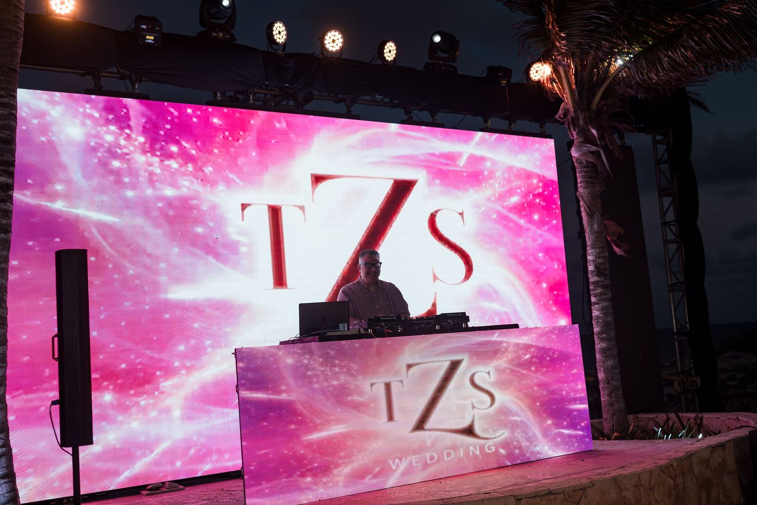 DJ performing on stage with a vibrant pink TZS logo display at an outdoor nighttime event.
