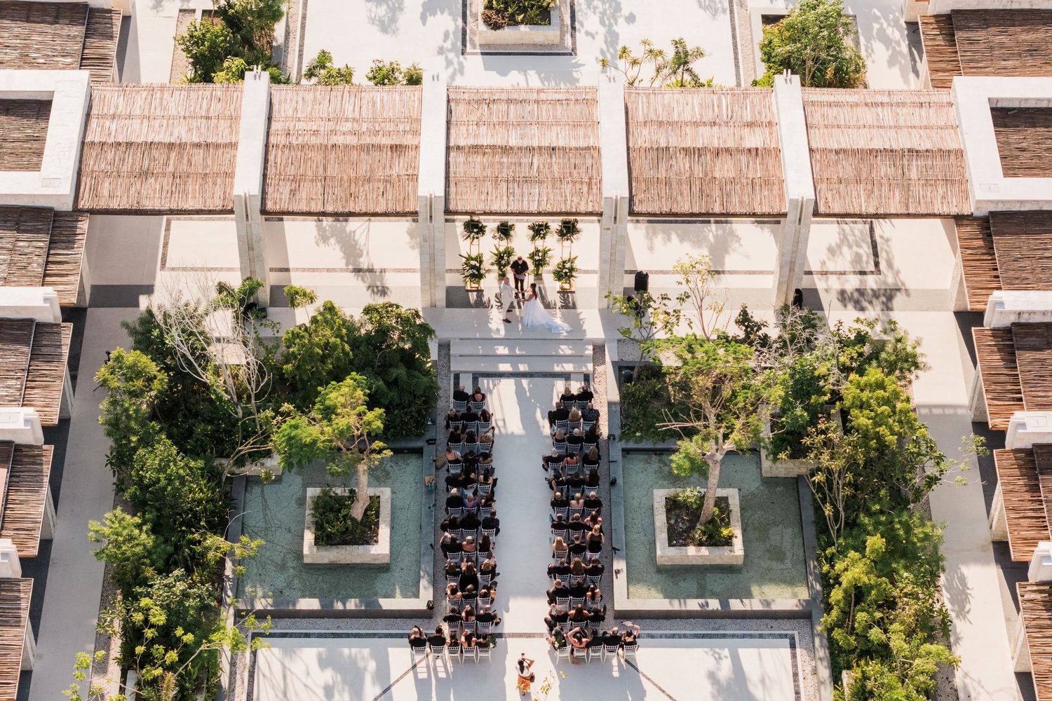 Aerial view of elegant outdoor wedding ceremony with guests seated in rows between lush tropical gardens.