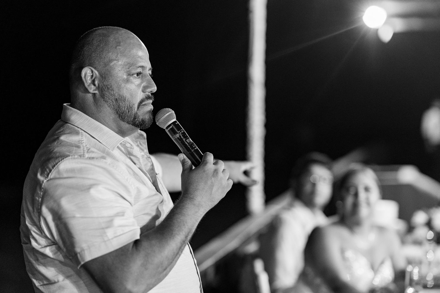 Man speaking into microphone at wedding reception in black and white photo with guests in background.