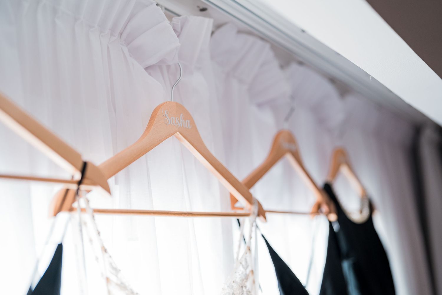 Wooden rose gold hangers holding white wedding veil and black bridesmaid dresses on a curtain rod.