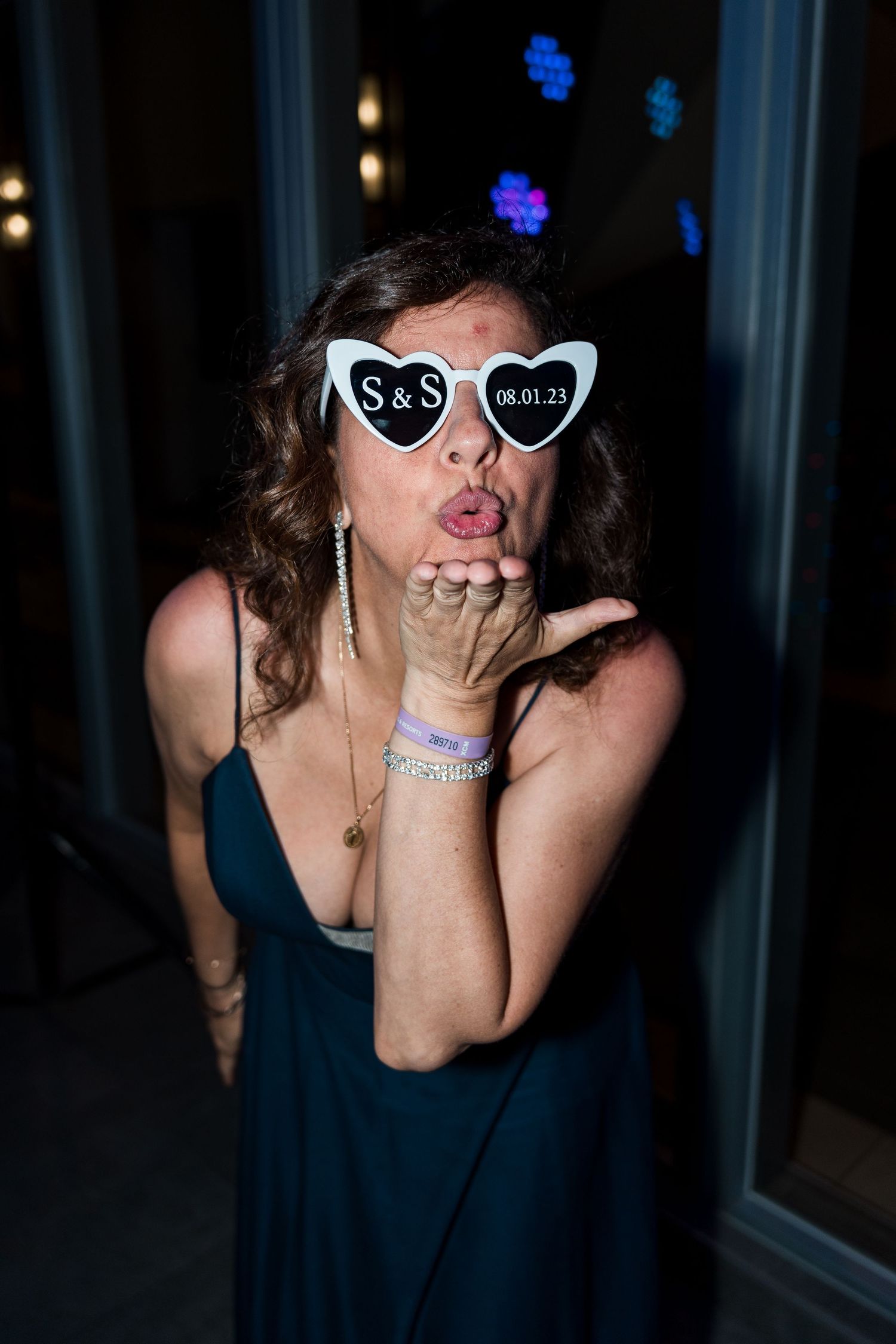 Woman wearing S&S heart-shaped wedding sunglasses blows a kiss at evening wedding reception.