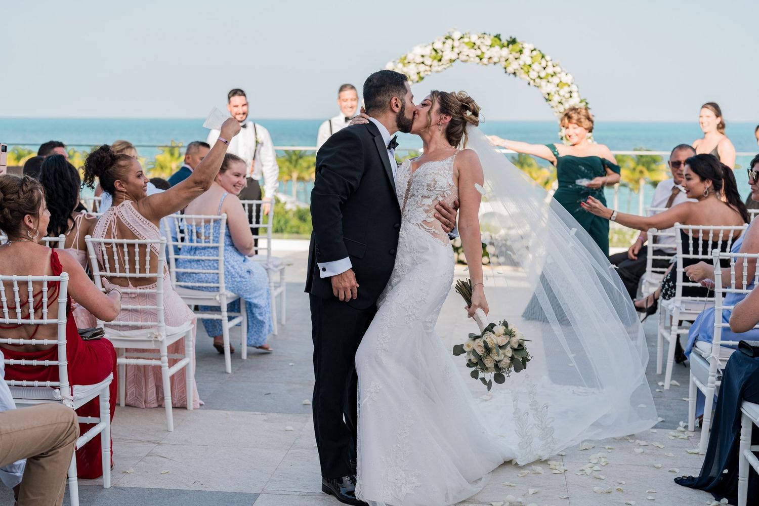 Bride and groom share first kiss at elegant outdoor beach wedding ceremony with floral arch.